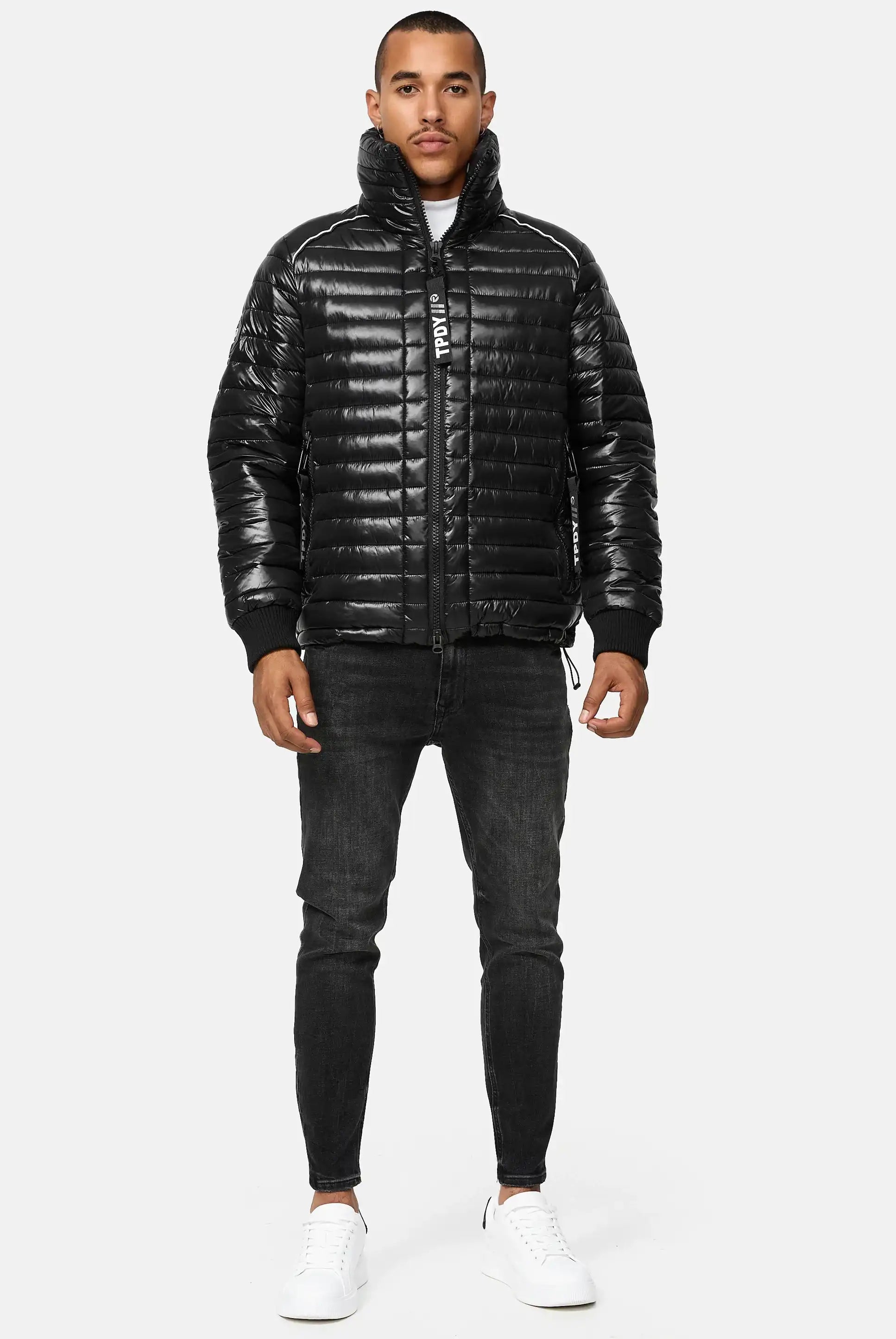 Ein Mann sieht nach vorne und trägt eine glänzende schwarze Corey Steppjacke mit Stehkragen, schwarze Skinny Jeans und weiße Turnschuhe vor einem schlichten hellgrauen Hintergrund.