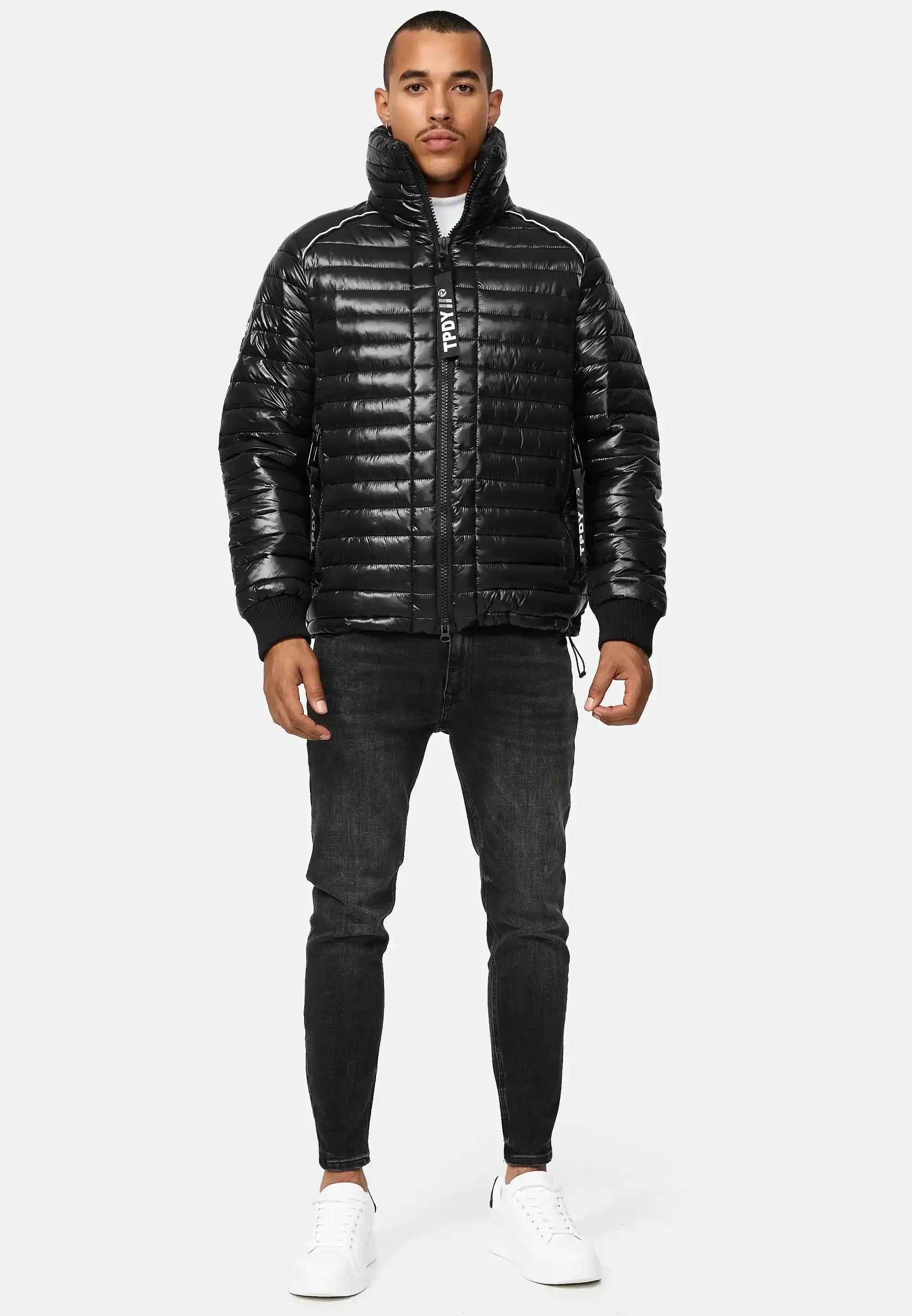 Ein Mann sieht nach vorne und trägt eine glänzende schwarze Corey Steppjacke mit Stehkragen, schwarze Skinny Jeans und weiße Turnschuhe vor einem schlichten hellgrauen Hintergrund.