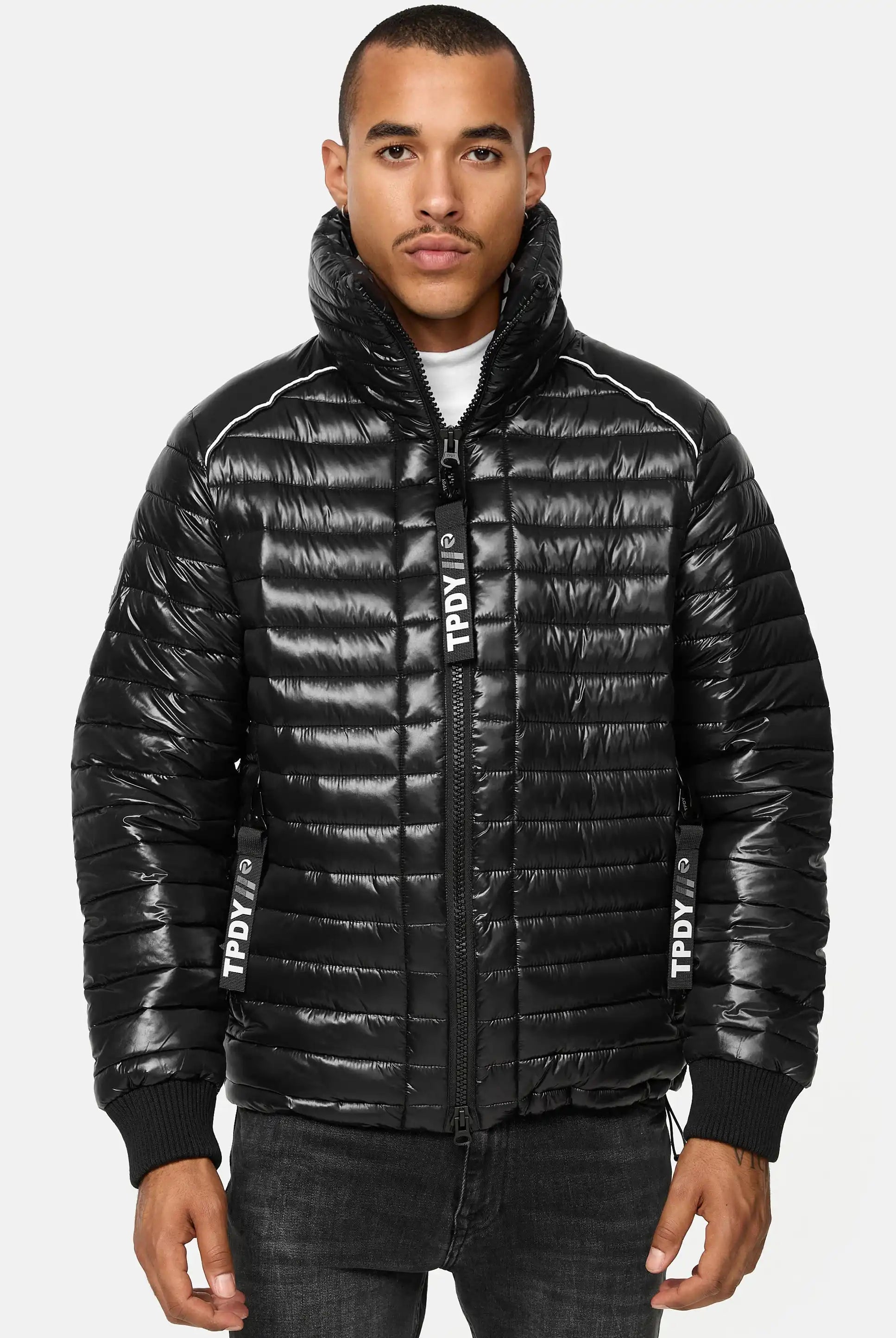Ein Mann steht mit dem Gesicht nach vorne und trägt eine glänzende schwarze Trueprodigy Corey Steppjacke mit Stehkragen mit TPDIV in Weiß auf der Vorderseite und dem Reißverschluss, gepaart mit einer dunklen Jeans. Die Jacke hat einen hohen Kragen und eine gesteppte Textur.