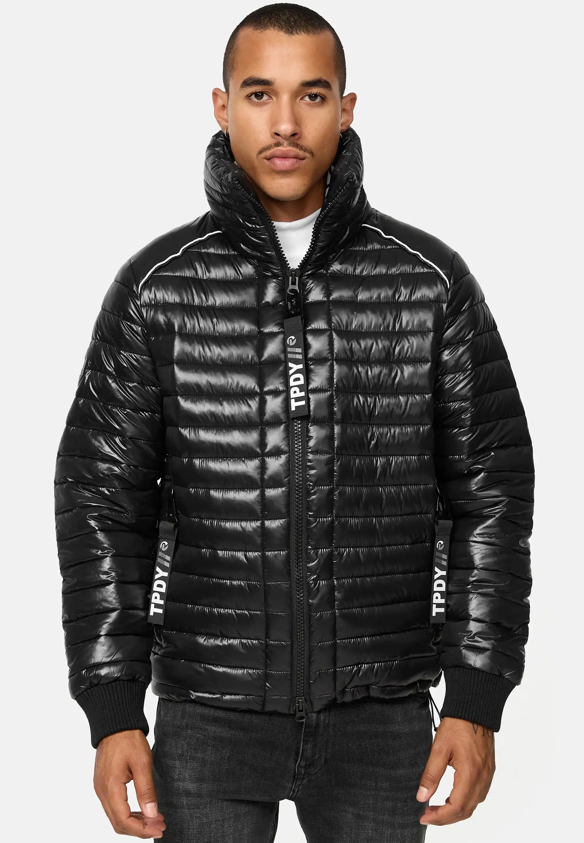 Ein Mann steht mit dem Gesicht nach vorne und trägt eine glänzende schwarze Trueprodigy Corey Steppjacke mit Stehkragen mit TPDIV in Weiß auf der Vorderseite und dem Reißverschluss, gepaart mit einer dunklen Jeans. Die Jacke hat einen hohen Kragen und eine gesteppte Textur.