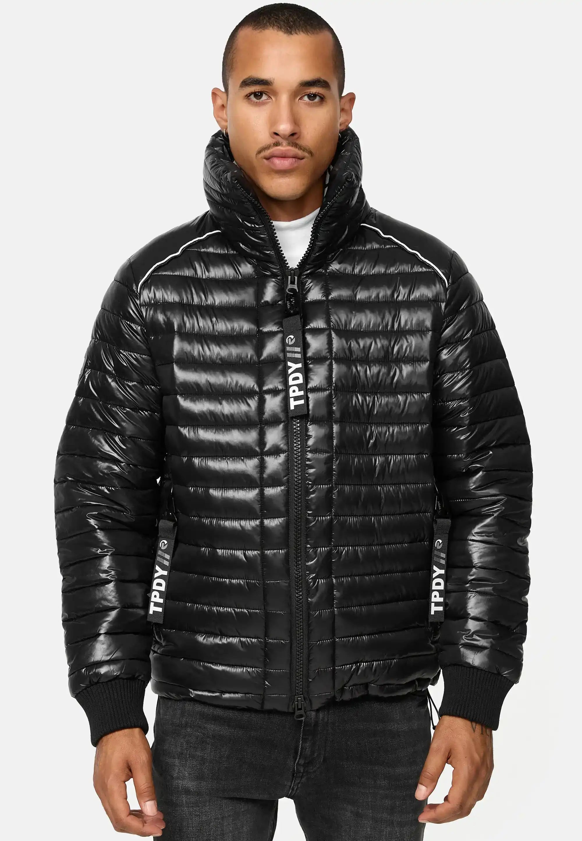 Ein Mann steht mit dem Gesicht nach vorne und trägt eine glänzende schwarze Trueprodigy Corey Steppjacke mit Stehkragen mit TPDIV in Weiß auf der Vorderseite und dem Reißverschluss, gepaart mit einer dunklen Jeans. Die Jacke hat einen hohen Kragen und eine gesteppte Textur.