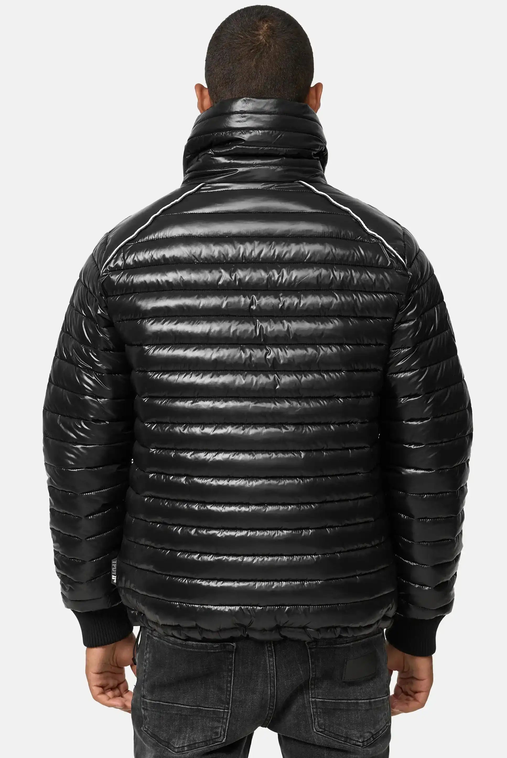 Eine Person ist von hinten zu sehen, die eine glänzende schwarze Trueprodigy Corey Steppjacke mit Stehkragen mit horizontaler Steppung, hohem Kragen und gerippten Bündchen trägt, gepaart mit schwarzen Jeans vor einem schlichten hellen Hintergrund.