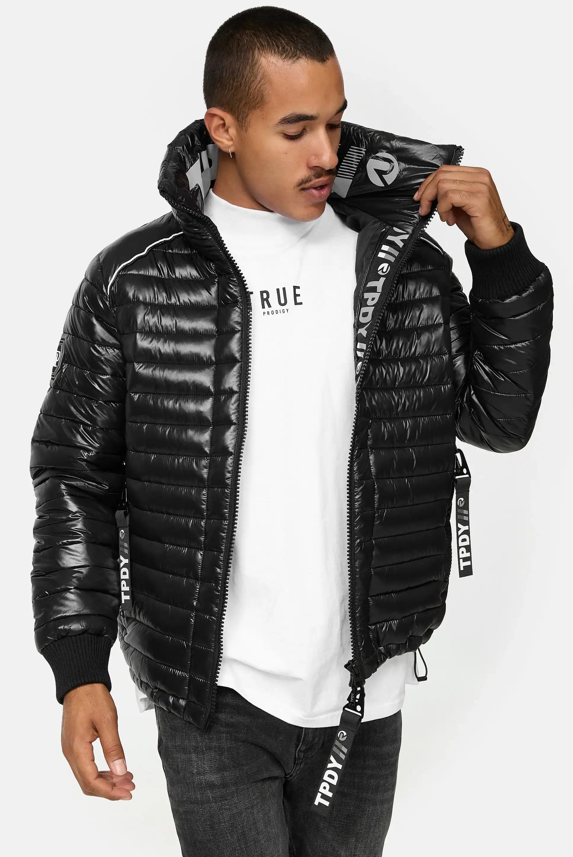 Ein Mann steht mit Blick nach vorne in einer glänzenden schwarzen Corey Steppjacke mit Stehkragen von trueprodigy über einem weißen T-Shirt und dunklen Jeans. Er öffnet den Reißverschluss der Jacke, schaut nach unten und hält sie mit einer Hand offen.