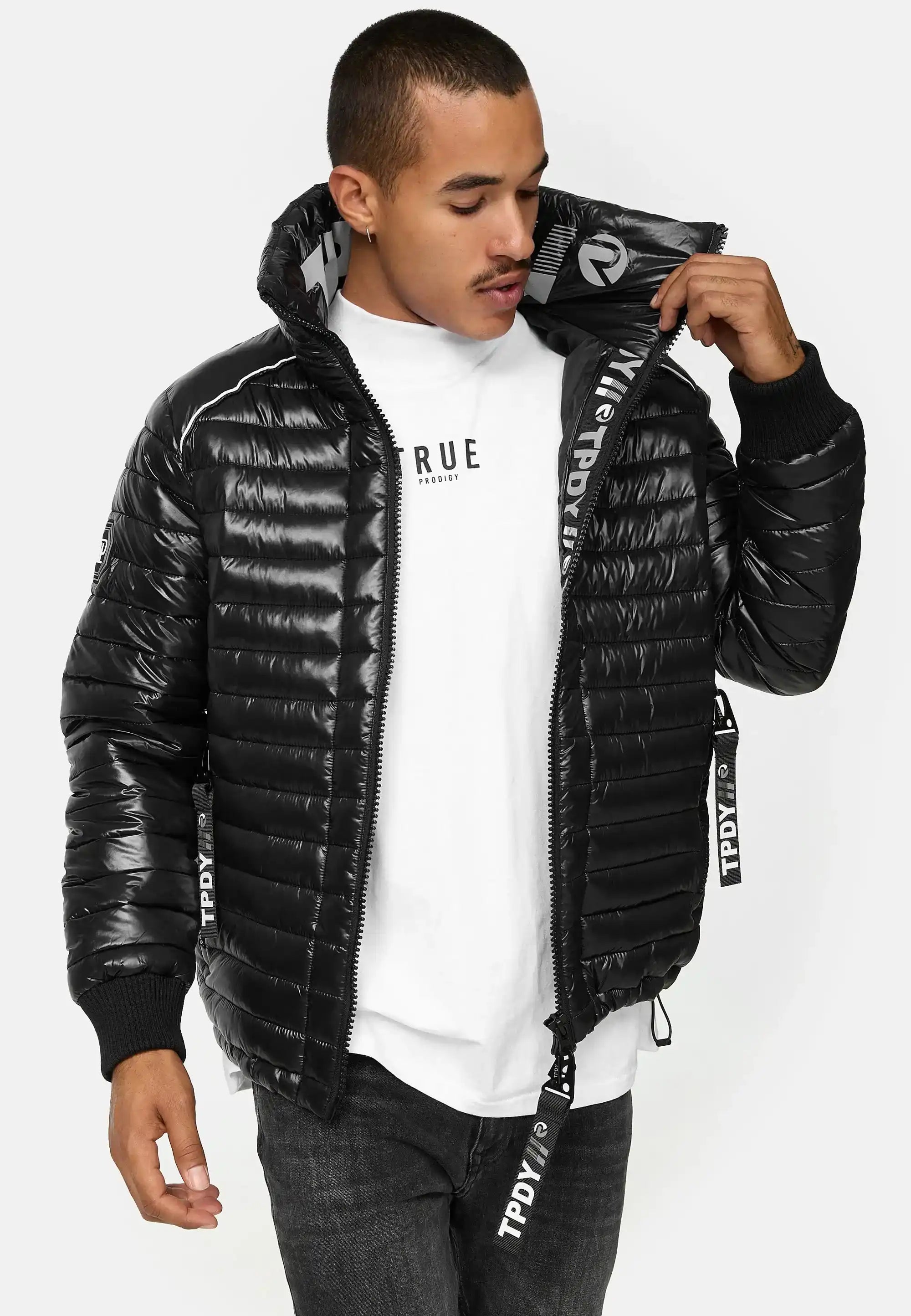 Ein Mann steht mit Blick nach vorne in einer glänzenden schwarzen Corey Steppjacke mit Stehkragen von trueprodigy über einem weißen T-Shirt und dunklen Jeans. Er öffnet den Reißverschluss der Jacke, schaut nach unten und hält sie mit einer Hand offen.