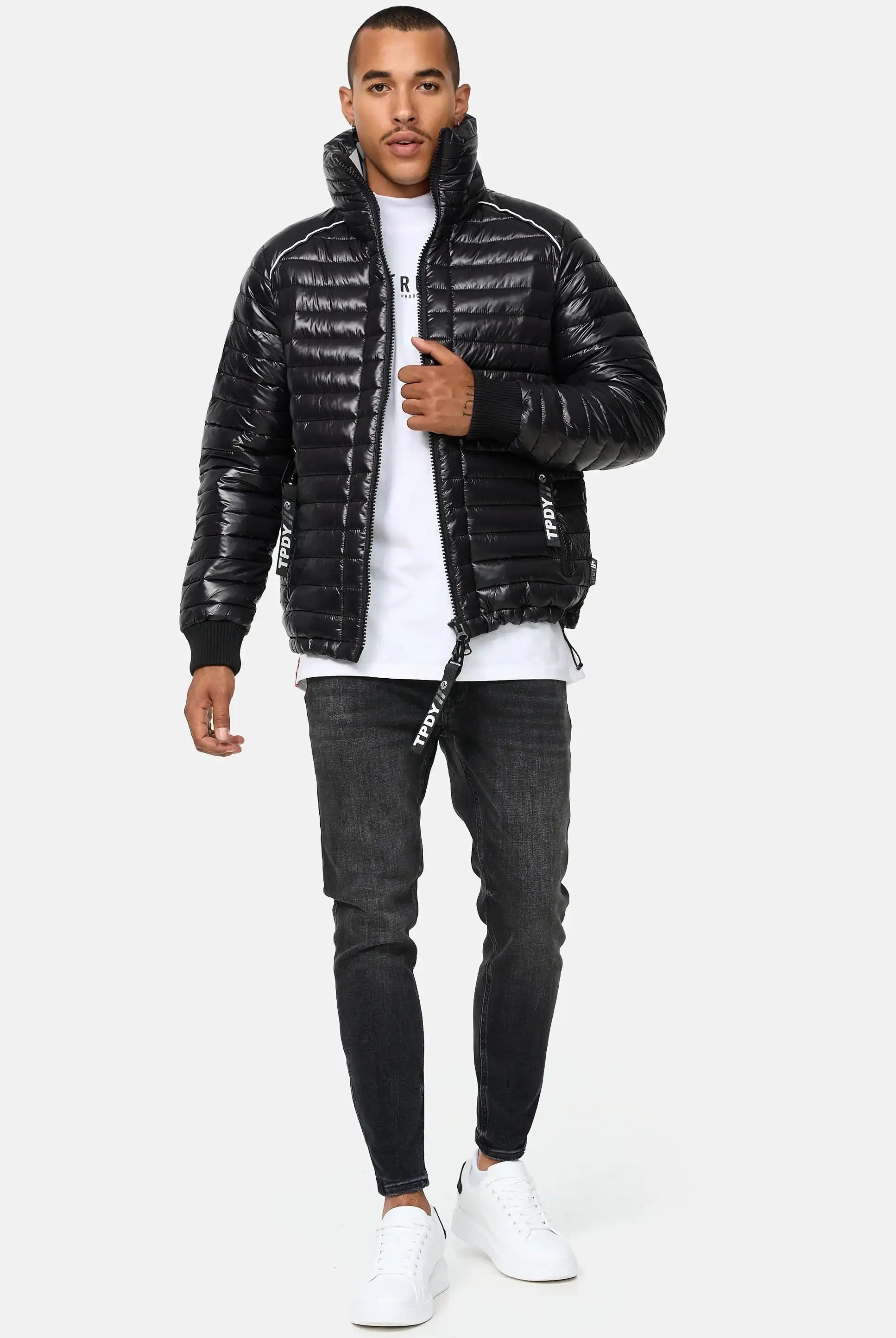 Ein Mann steht mit dem Gesicht nach vorne und trägt die Trueprodigy Corey Steppjacke mit Stehkragen, gepaart mit einem weißen T-Shirt, dunklen Skinny Jeans und weißen Turnschuhen. Eine Hand steckt in seiner Tasche, die andere hält den Reißverschluss der Jacke vor einem hellen Hintergrund.