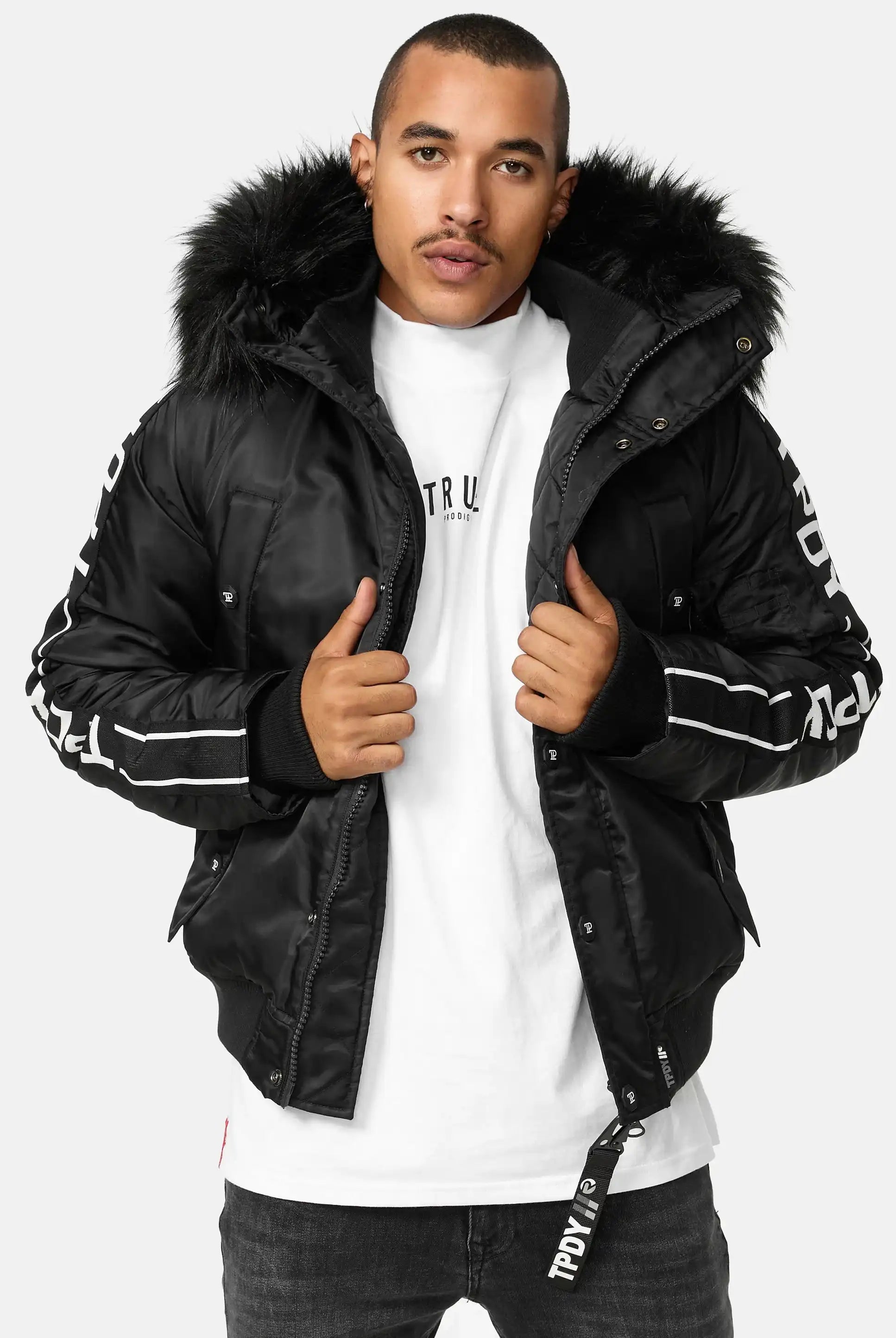 Ein Mann, der die trueprodigy Jonathan Bomberjacke mit Kapuze und Kunstfell, ein weißes T-Shirt und eine dunkle Jeans trägt, steht vor der Kamera und hält seine Jacke offen.