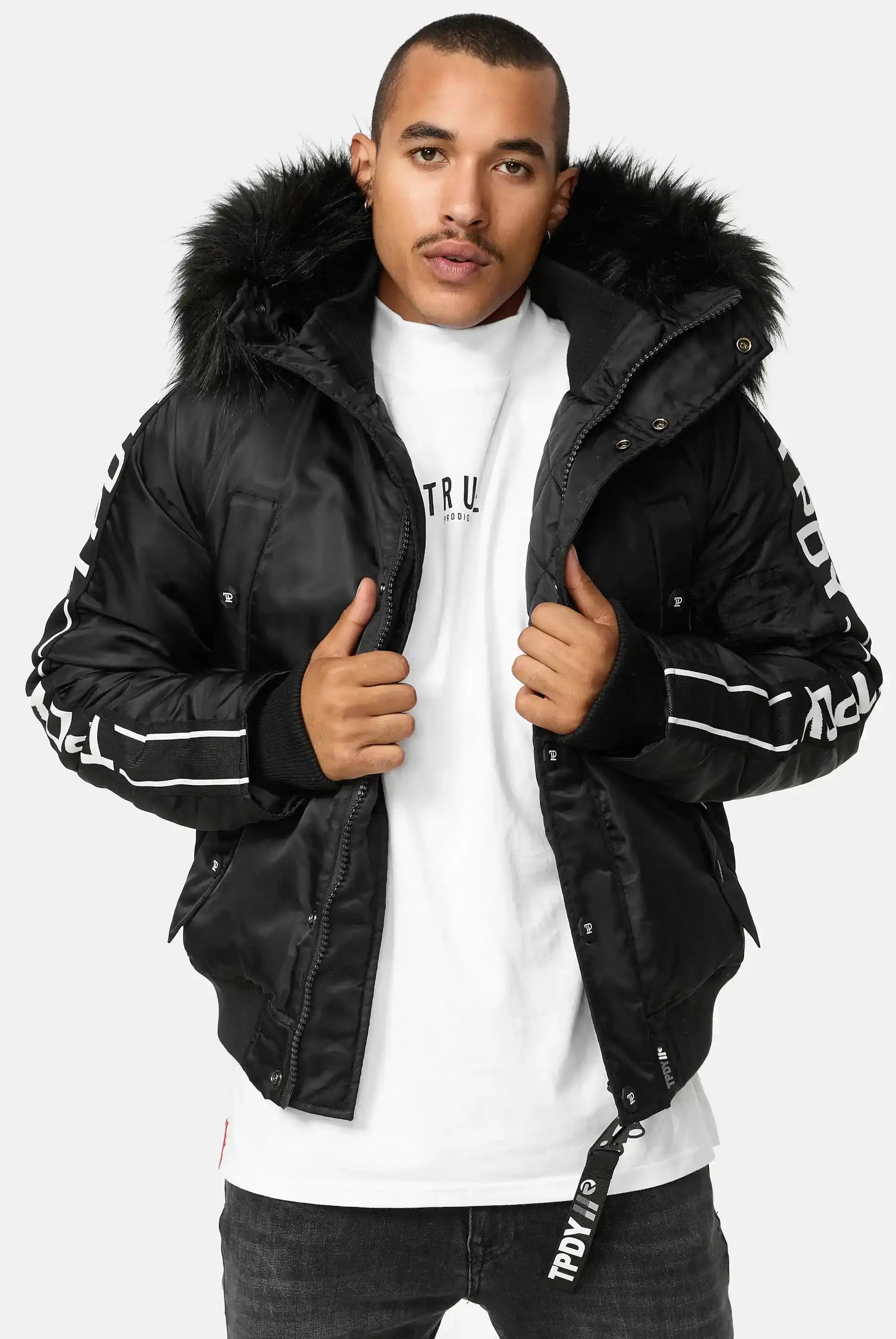 Ein Mann, der die trueprodigy Jonathan Bomberjacke mit Kapuze und Kunstfell, ein weißes T-Shirt und eine dunkle Jeans trägt, steht vor der Kamera und hält seine Jacke offen.