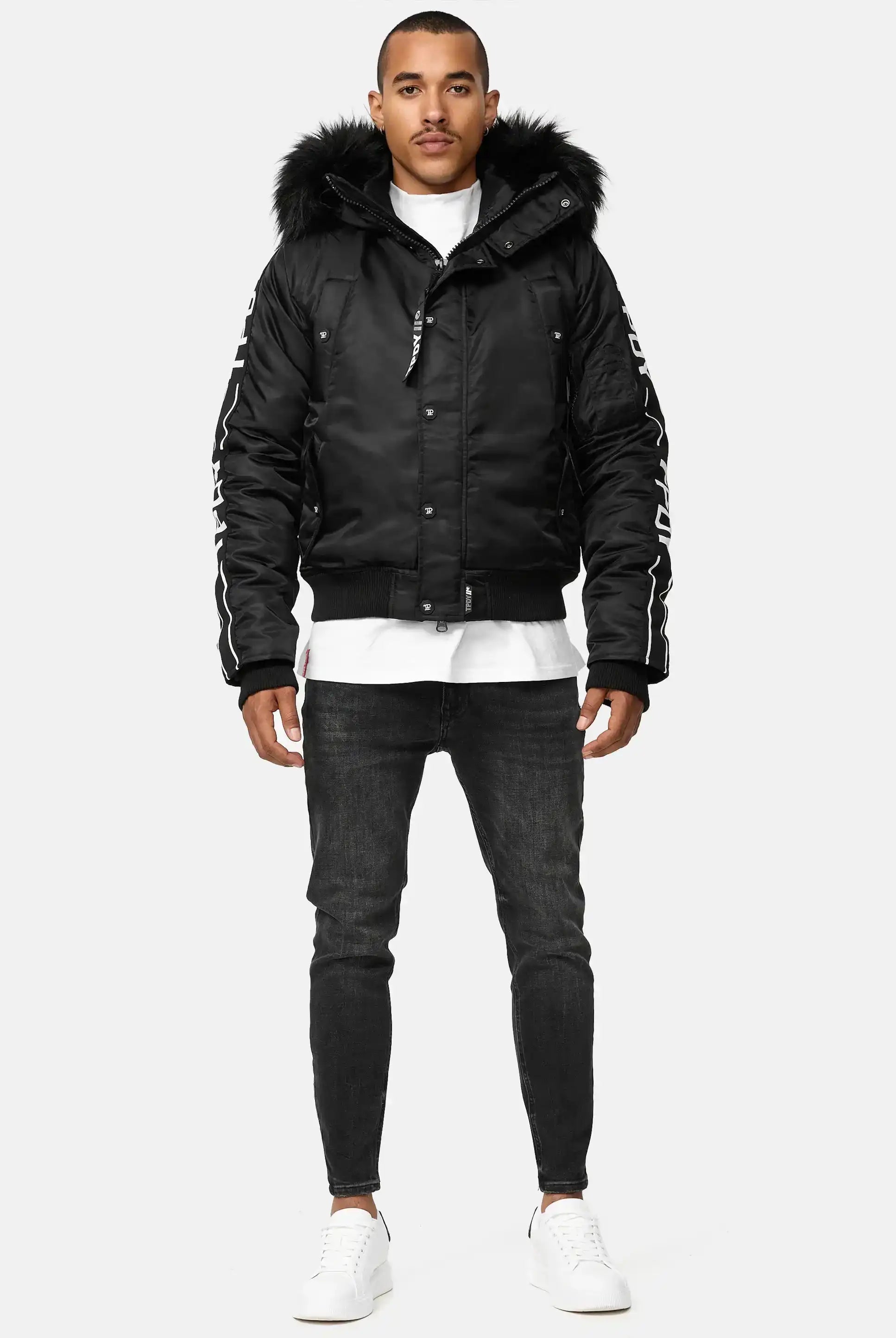Ein Mann trägt die Jonathan Bomberjacke mit Kapuze und Kunstfell von trueprodigy, kombiniert mit schwarzen Jeans und weißen Sneakern. Er steht frontal vor einem hellen Hintergrund, hat kurze Haare und einen gepflegten Bart.