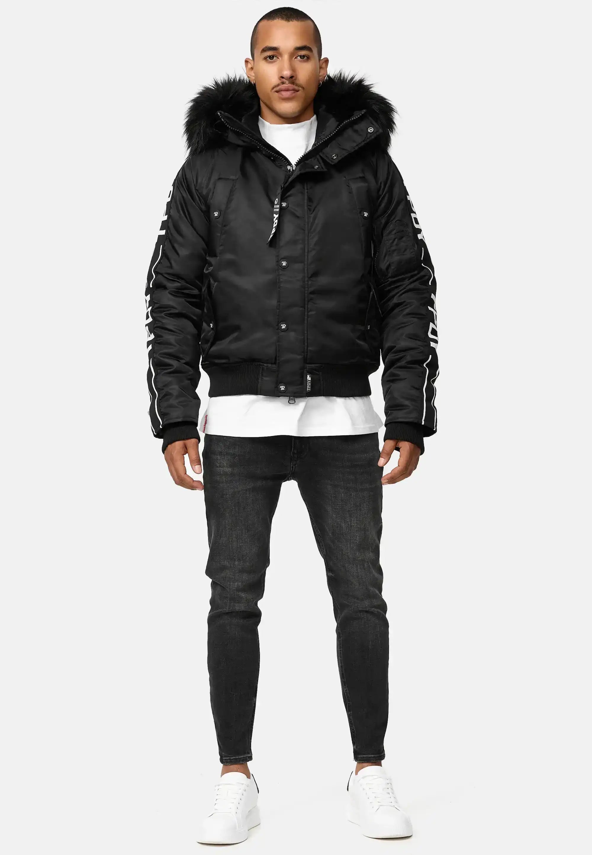 Ein Mann trägt die Jonathan Bomberjacke mit Kapuze und Kunstfell von trueprodigy, kombiniert mit schwarzen Jeans und weißen Sneakern. Er steht frontal vor einem hellen Hintergrund, hat kurze Haare und einen gepflegten Bart.