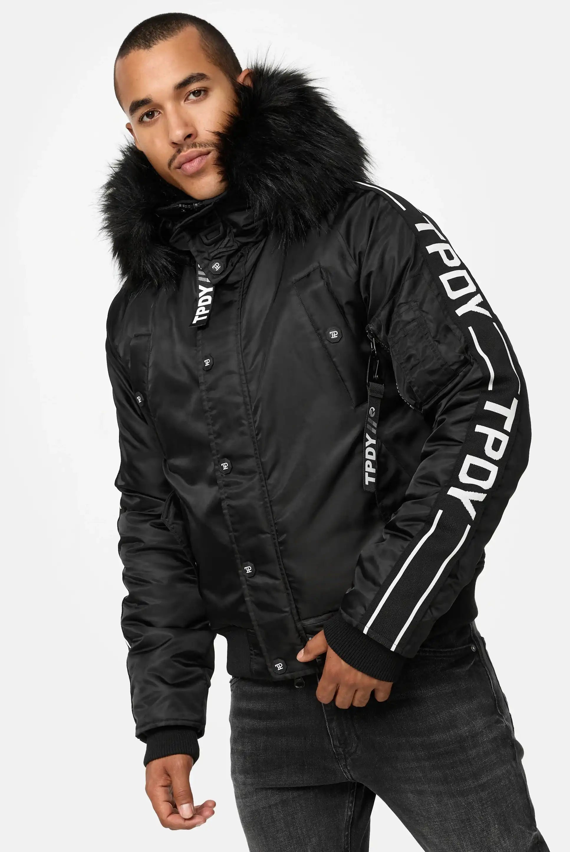 Ein Mann trägt die Jonathan Bomberjacke mit Kapuze und Kunstfell von trueprodigy sowie schwarze Jeans und steht vor einem schlichten weißen Hintergrund.