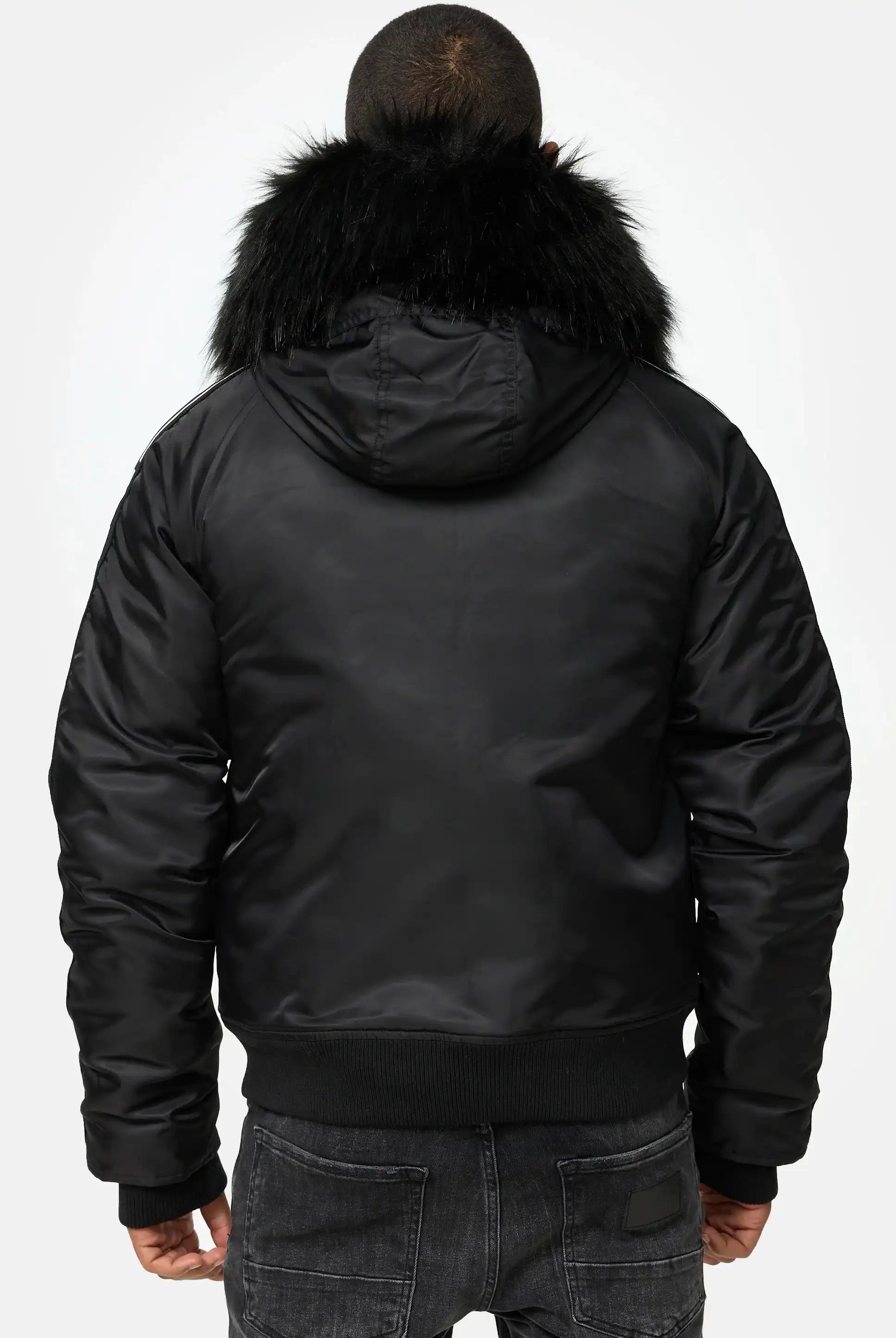 Eine Person steht mit dem Rücken zur Kamera und trägt die trueprodigy Jonathan Bomberjacke mit Kapuze und Kunstfell und dunkle Jeans. Der Hintergrund ist einfarbig und hell.