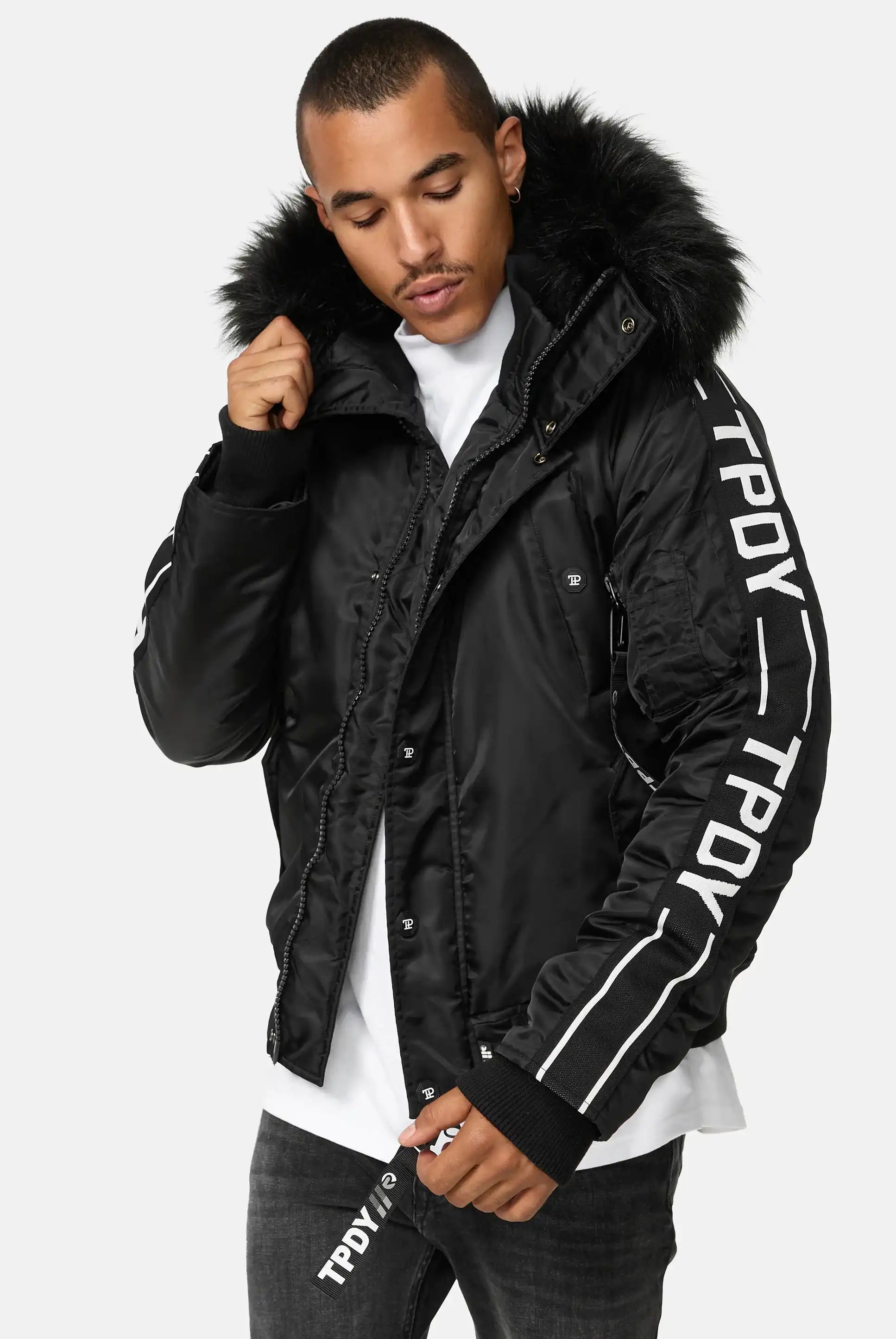 Eine Person, die die Jonathan Bomberjacke mit Kapuze und Kunstfell von trueprodigy mit dem Text TPDY auf den Ärmeln und dem Reißverschluss trägt, über einem weißen Hemd und einer dunklen Jeans, steht vor einem schlichten hellen Hintergrund.