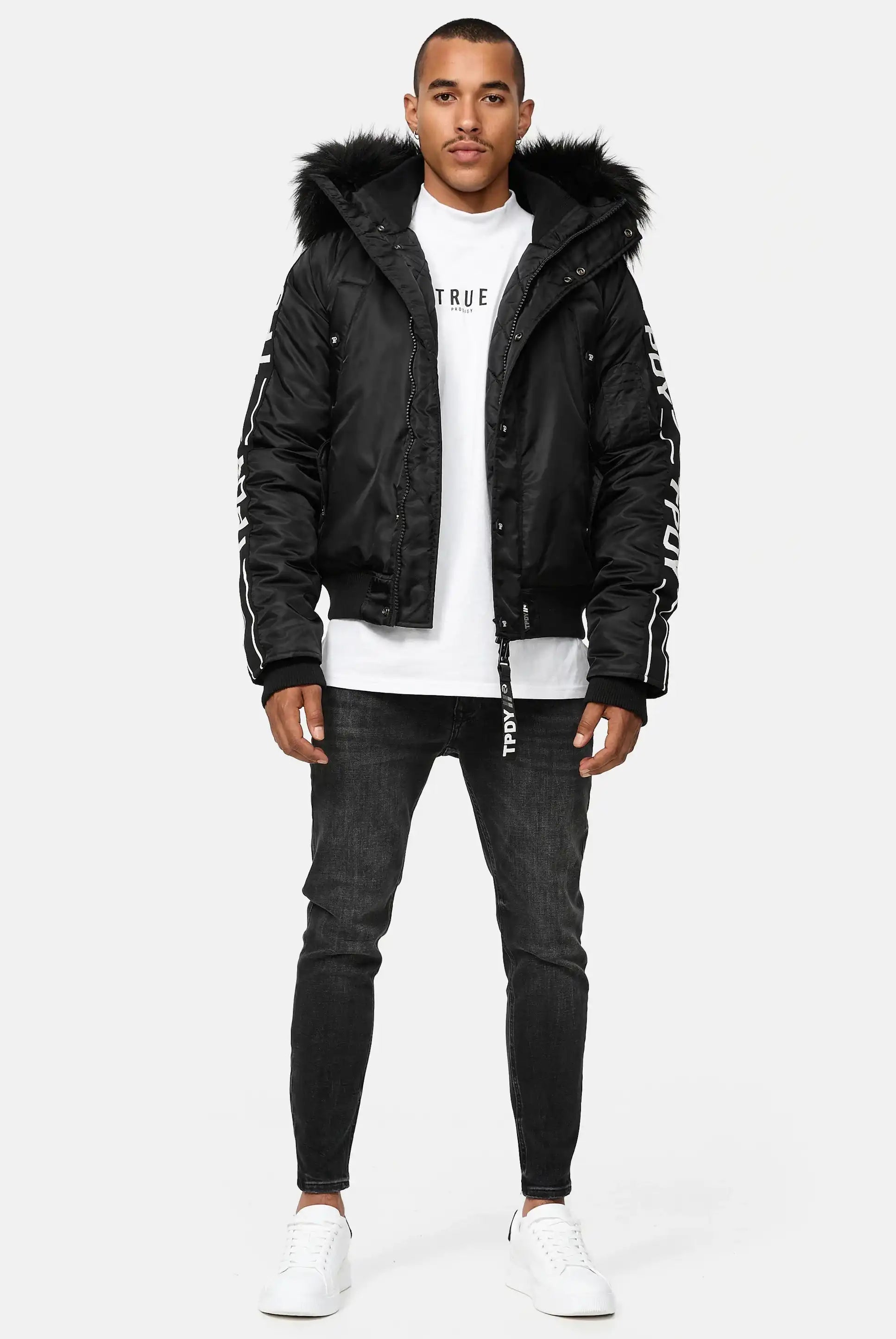 Ein Mann trägt die Jonathan Bomberjacke mit Kapuze und Kunstfell von trueprodigy, dazu ein weißes Shirt, schwarze Skinny Jeans und weiße Sneaker vor schlichtem weißen Hintergrund.