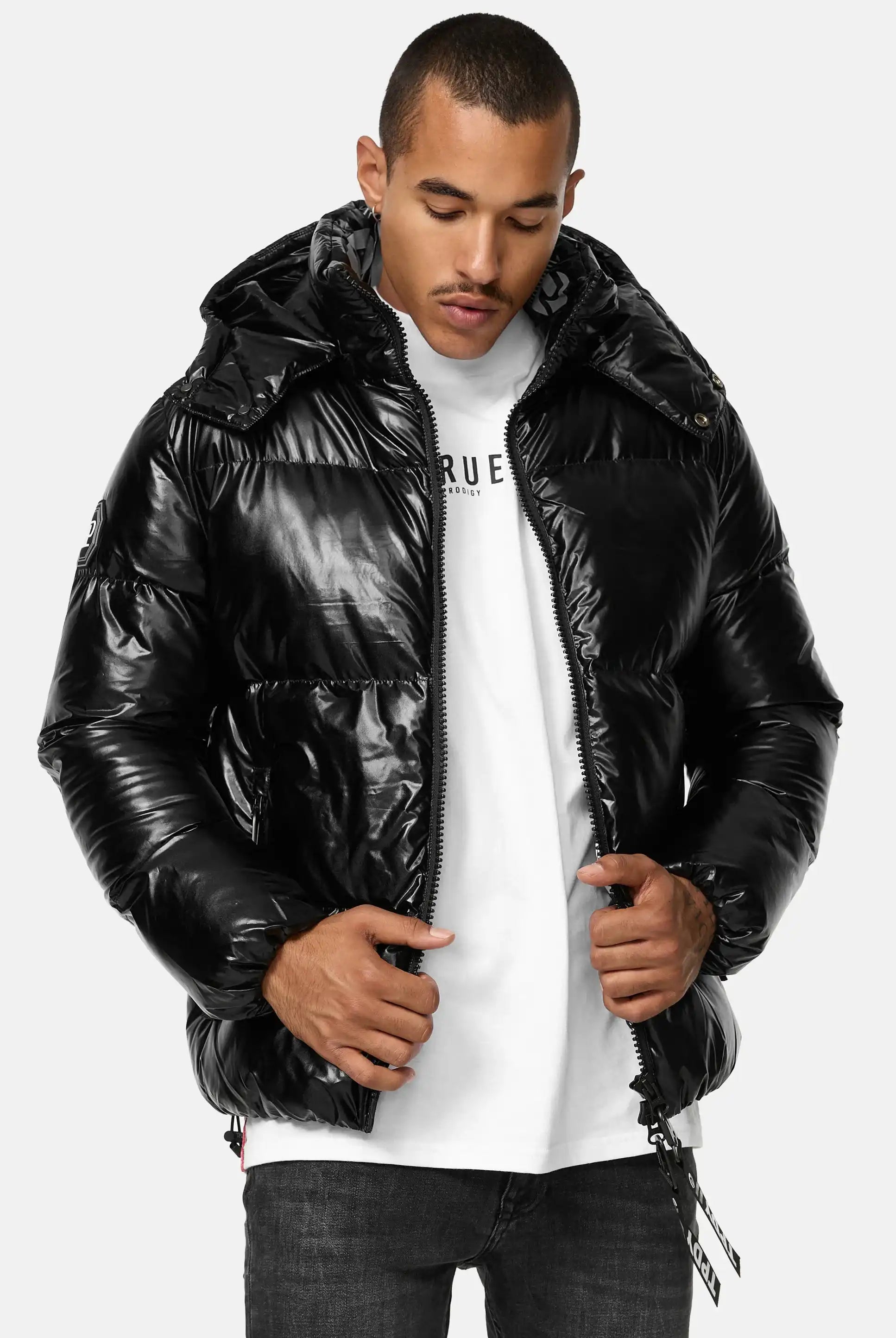 Mann trägt glänzende schwarze TRUEPRODIGY constance Pufferjacke mit Kapuze, darunter weißes T-Shirt mit „TRUEPRODIGY“-Logo, kombiniert mit dunkelgrauer Jeans. Ideal für stilvolle Wintermode.