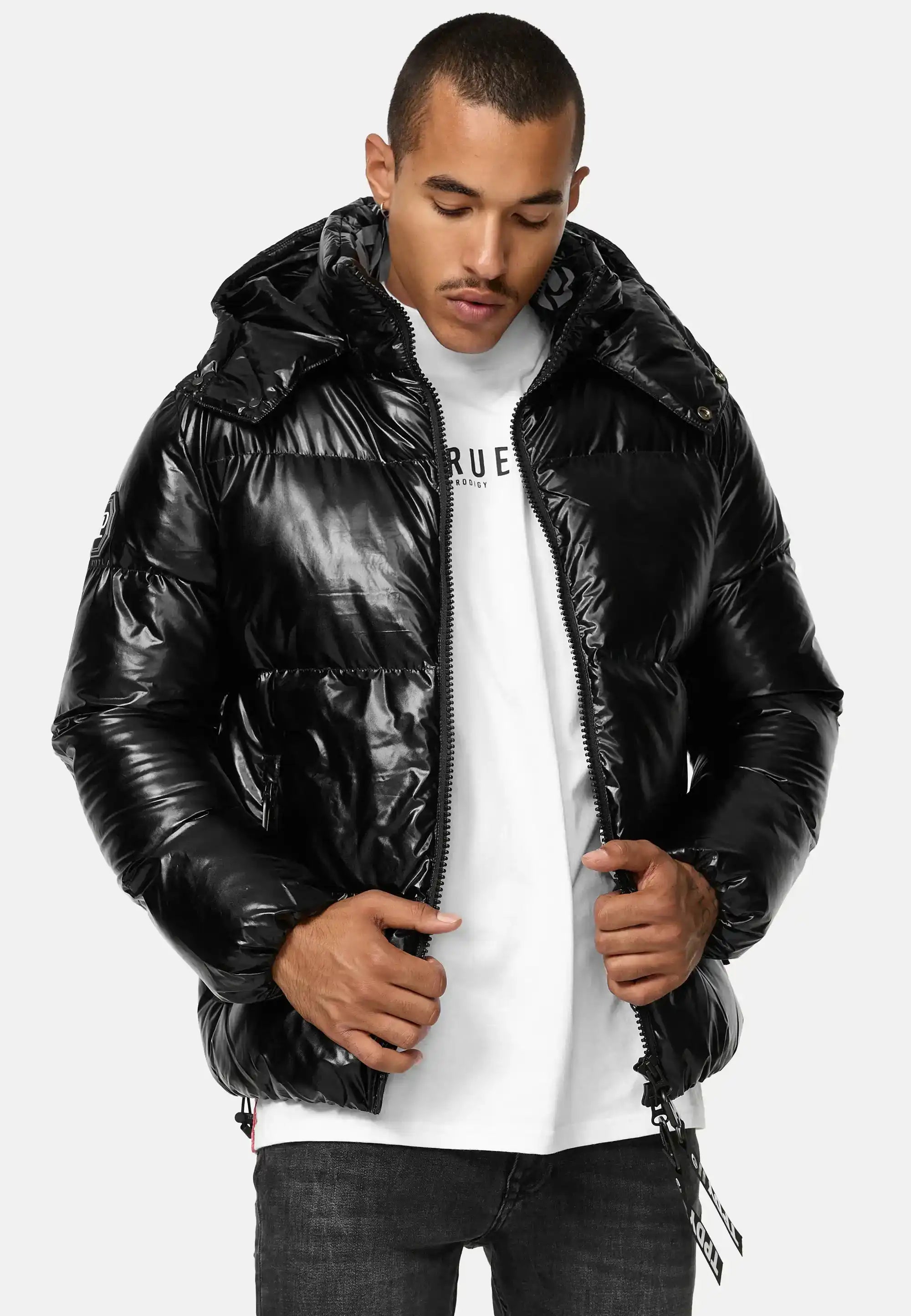 Mann trägt glänzende schwarze TRUEPRODIGY constance Pufferjacke mit Kapuze, darunter weißes T-Shirt mit „TRUEPRODIGY“-Logo, kombiniert mit dunkelgrauer Jeans. Ideal für stilvolle Wintermode.