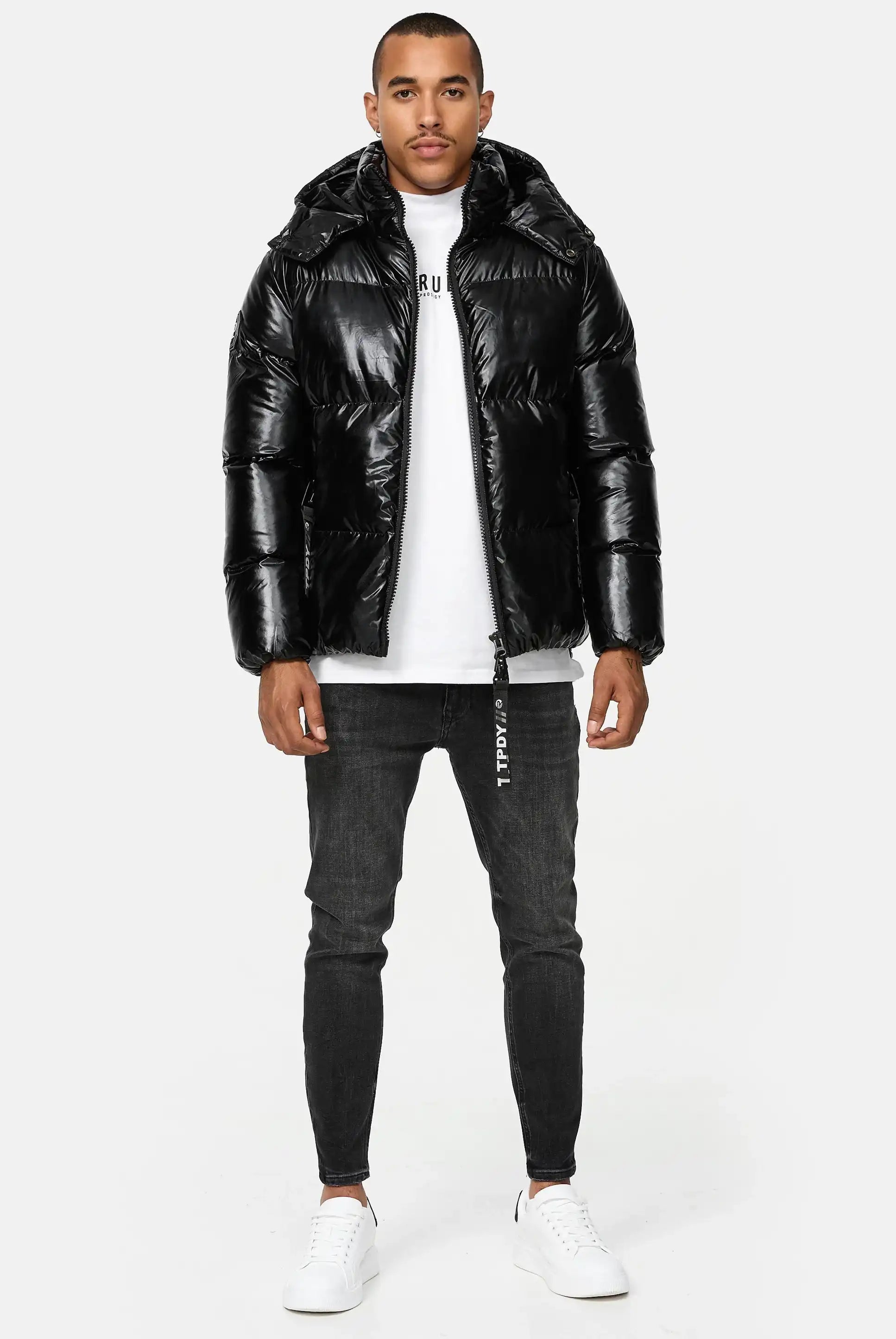 Mann trägt schwarze TRUEPRODIGY constance Pufferjacke mit glänzendem Finish, weißes T-Shirt mit „TRUE“ Schriftzug, schwarze Skinny Jeans, weiße Sneaker. Mode, urbaner Stil, lässiges Design, wintertauglich.