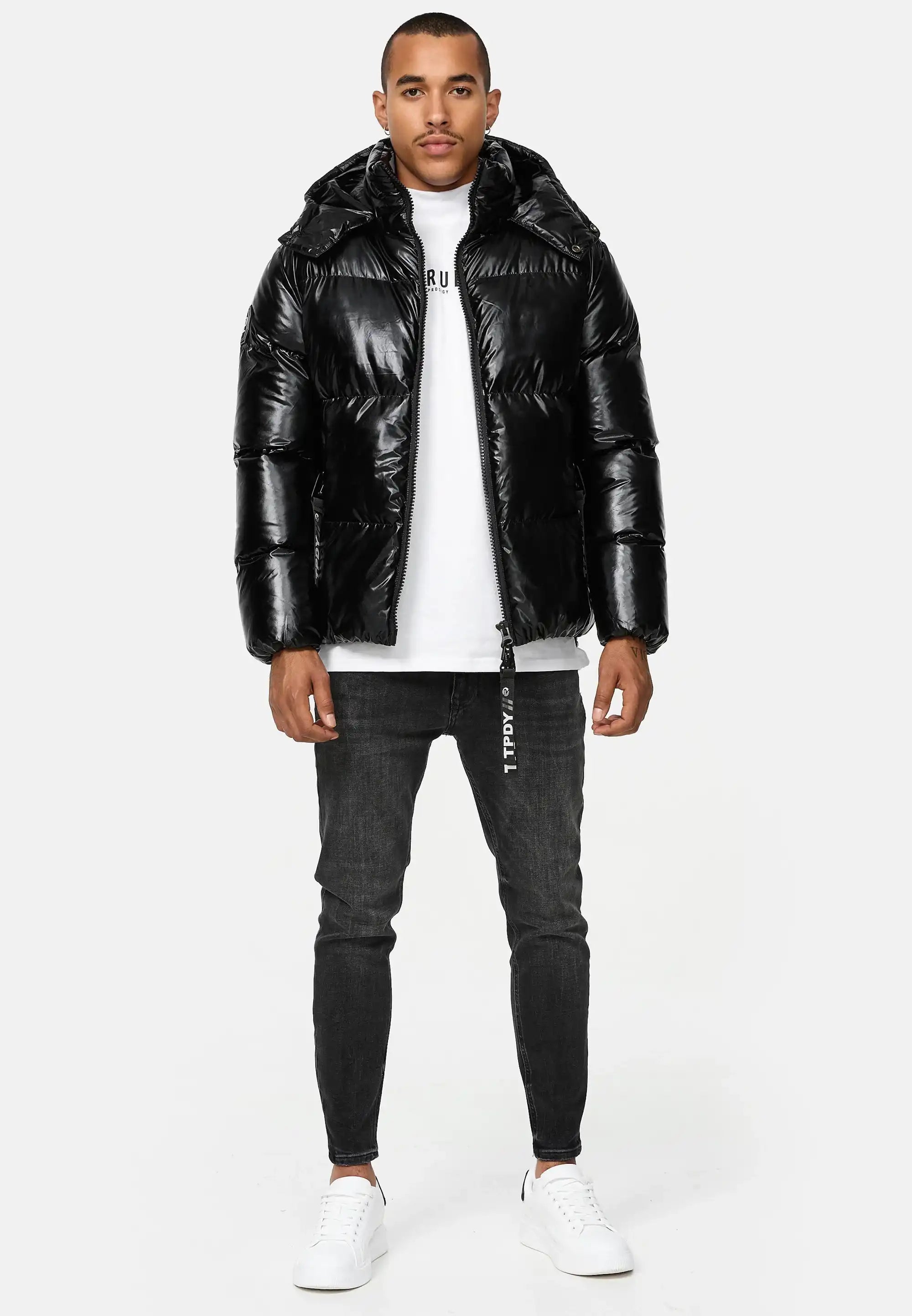 Mann trägt schwarze TRUEPRODIGY constance Pufferjacke mit glänzendem Finish, weißes T-Shirt mit „TRUE“ Schriftzug, schwarze Skinny Jeans, weiße Sneaker. Mode, urbaner Stil, lässiges Design, wintertauglich.