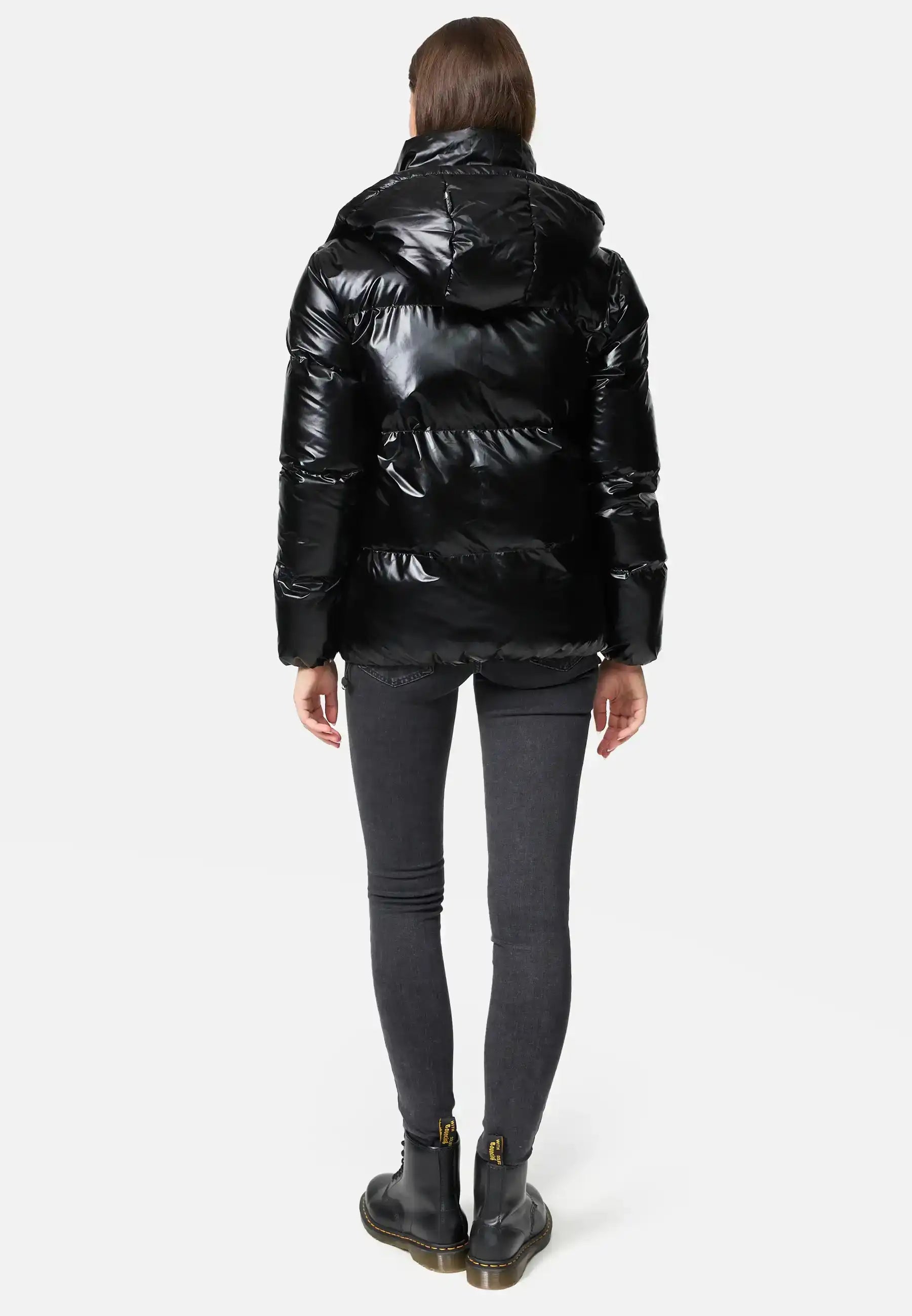 Frau trägt TRUEPRODIGY constance schwarze Steppjacke, Schriftzug „TRUEDPDE“, kombiniert mit schwarzen Jeans und Stiefeln. Jacke glänzend, elegant, wintertauglich, urbaner Stil.