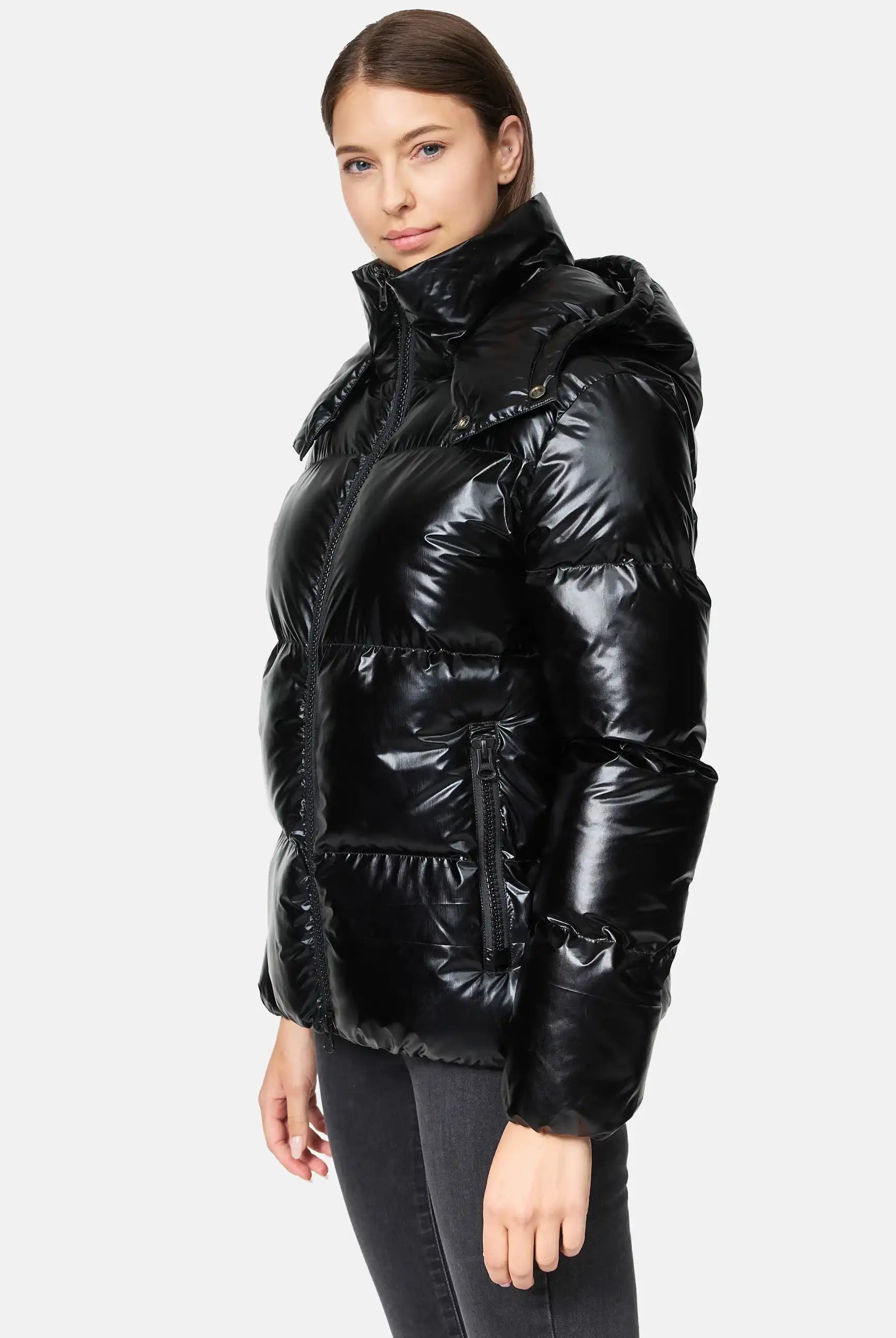 Eine Person in einer glänzenden schwarzen trueprodigy Constance F Glänzende Winterjacke mit Kapuze, die einen hohen Kragen und Reißverschlusstaschen aufweist, steht seitlich und blickt in die Kamera vor einem schlichten hellen Hintergrund.