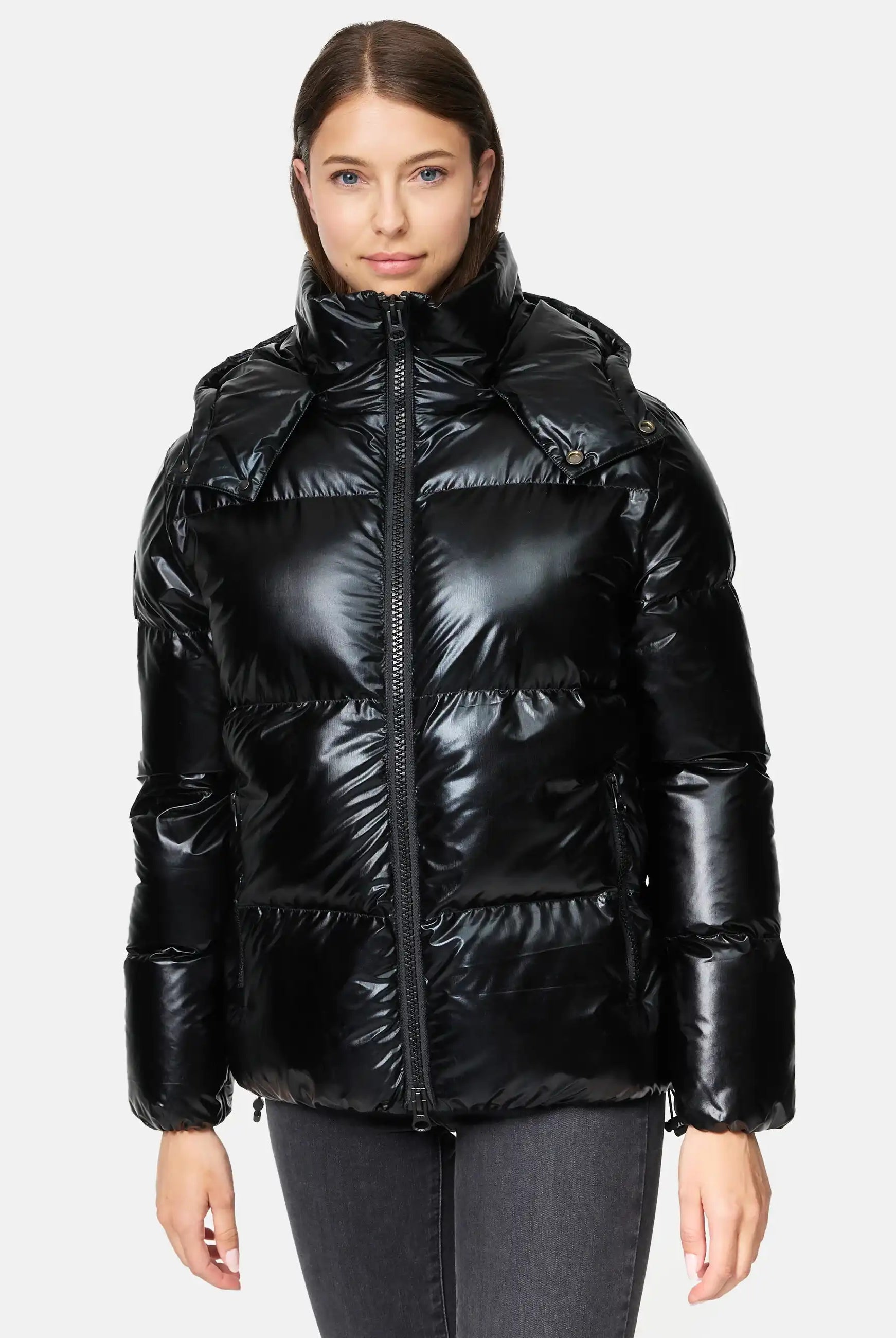 Eine Person steht mit dem Gesicht nach vorne und trägt die trueprodigy Constance F Glänzende Winterjacke mit Kapuze - eine glänzende schwarze Puffjacke mit hohem Kragen und Kapuze, die mit einer dunkelgrauen Jeans vor einem schlichten, hellen Hintergrund kombiniert wird.