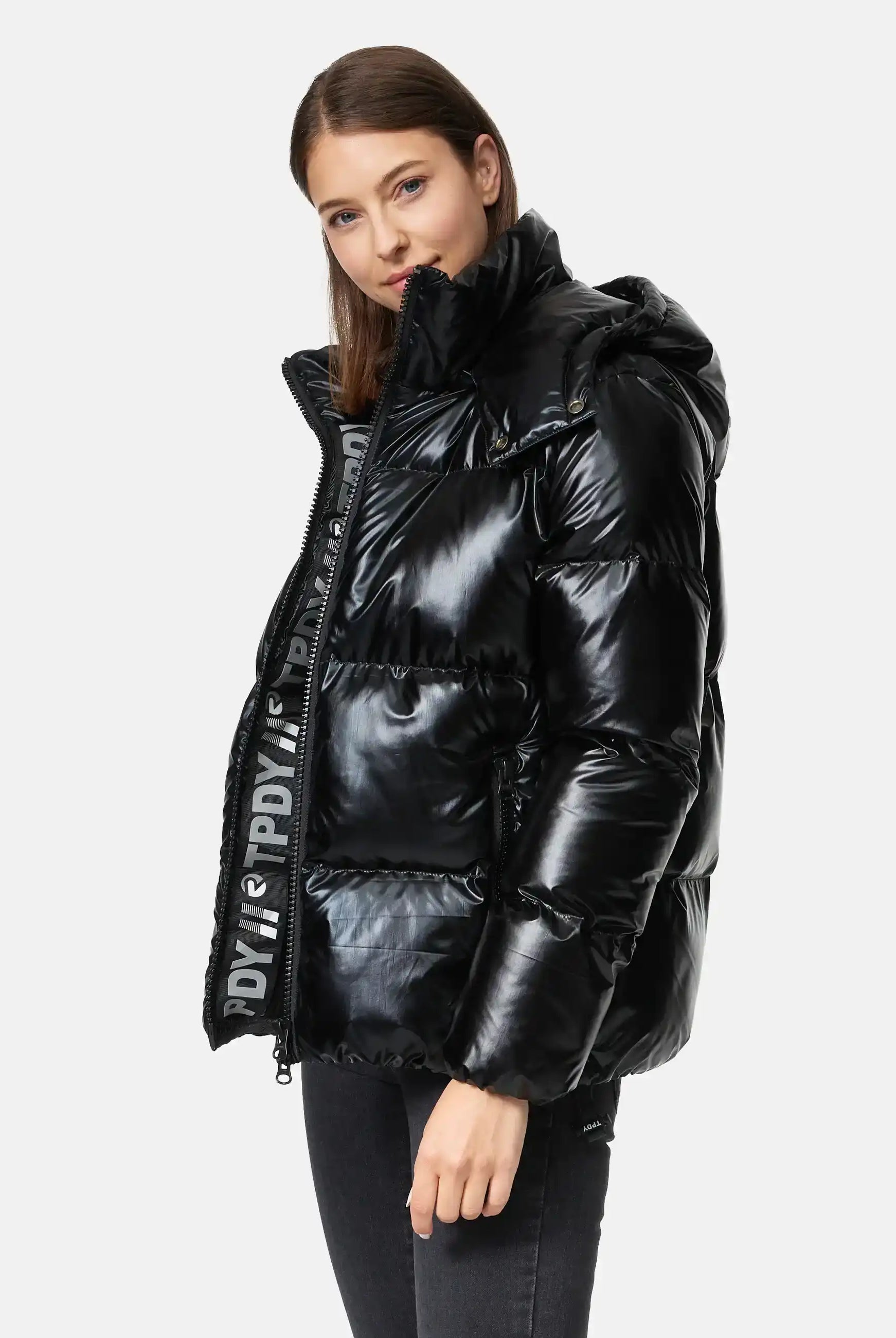 Eine Frau trägt die trueprodigy Constance F Glänzende Winterjacke mit Kapuze in glänzendem Schwarz mit hohem Kragen und gebrandetem Innenfutter, gepaart mit schwarzen Jeans, vor einem schlichten weißen Hintergrund.