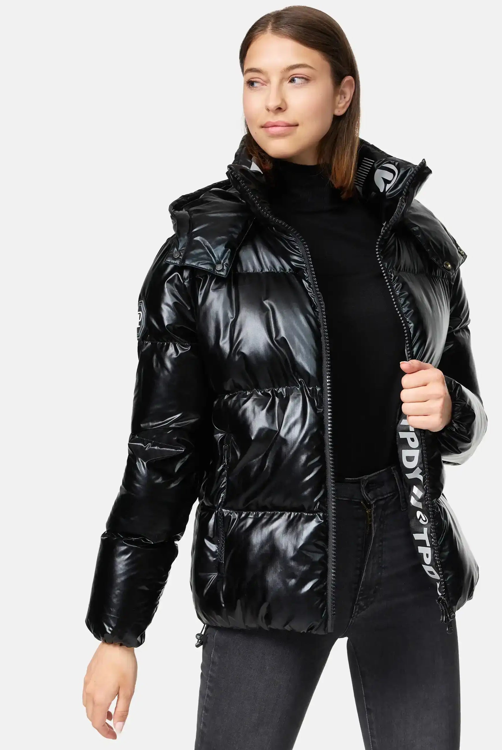 Eine Frau trägt die trueprodigy Constance F Glänzende Winterjacke mit Kapuze über einem schwarzen Rollkragenpullover und einer dunkelgrauen Jeans. Sie zieht den Reißverschluss der glänzenden Kapuzenjacke hoch und blickt mit einem neutralen Blick zur Seite.
