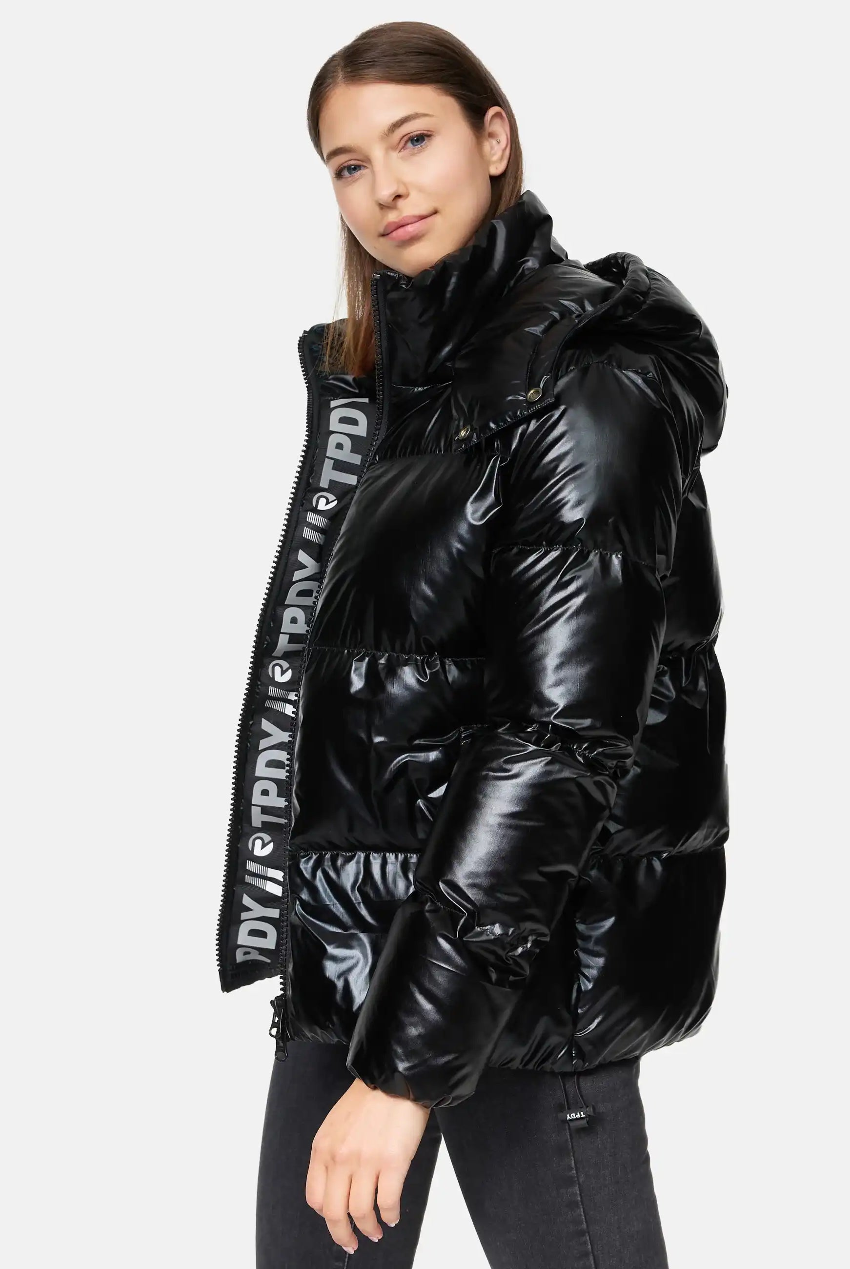 Eine Frau steht vor einem unifarbenen Hintergrund und trägt die trueprodigy Constance F Glänzende Winterjacke mit Kapuze, die einen großen Kragen und einen sichtbaren Reißverschluss mit aufgedrucktem Text aufweist, gepaart mit einer eleganten schwarzen Jeans.