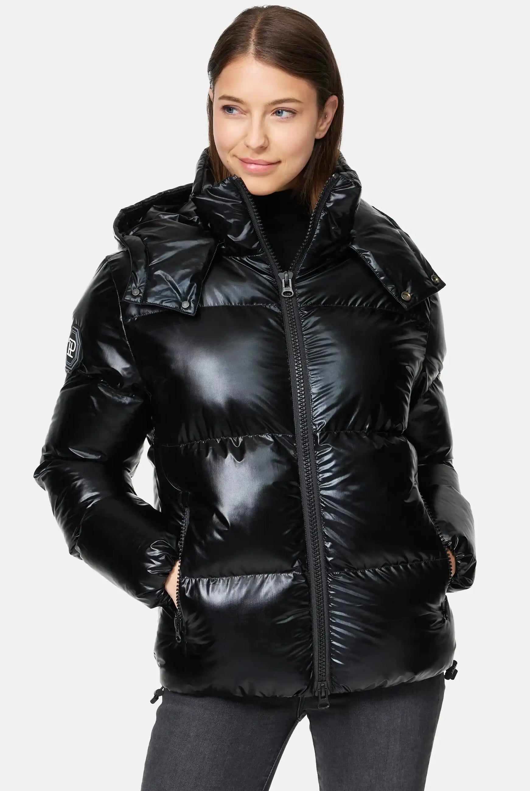 Frau trägt TRUEPRODIGY constance schwarze, glänzende, gesteppte Pufferjacke mit hoher Kapuze; seitliche Taschen, markantes Logo am Ärmel, ideal für Wintermode. Perfekte Kombination aus Stil und Wärme.