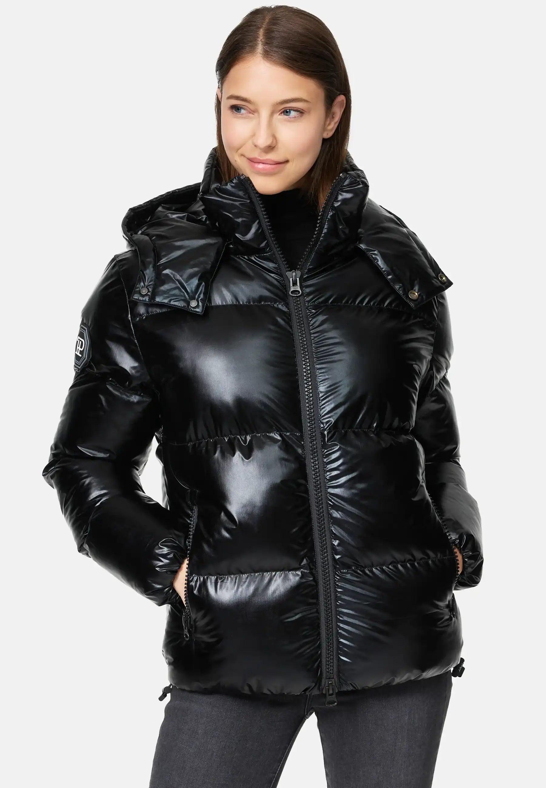 Frau trägt TRUEPRODIGY constance schwarze, glänzende, gesteppte Pufferjacke mit hoher Kapuze; seitliche Taschen, markantes Logo am Ärmel, ideal für Wintermode. Perfekte Kombination aus Stil und Wärme.