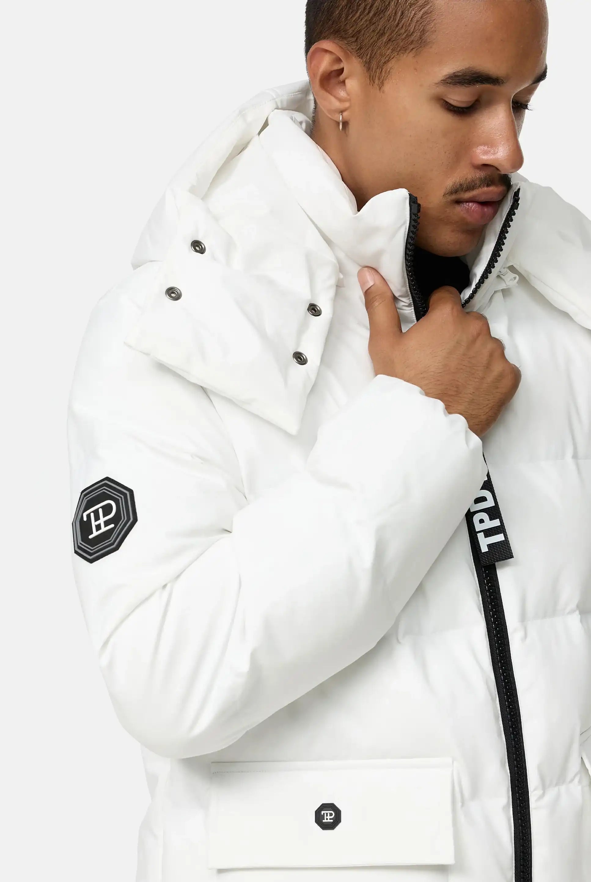 Eine Person in der Trueprodigy Franco Winterjacke mit abnehmbarer Kapuze, weiß mit schwarzen Reißverschlüssen und Logo-Patches an Ärmel und Tasche, steht vor hellem Hintergrund, blickt nach unten und hält den Kragen.