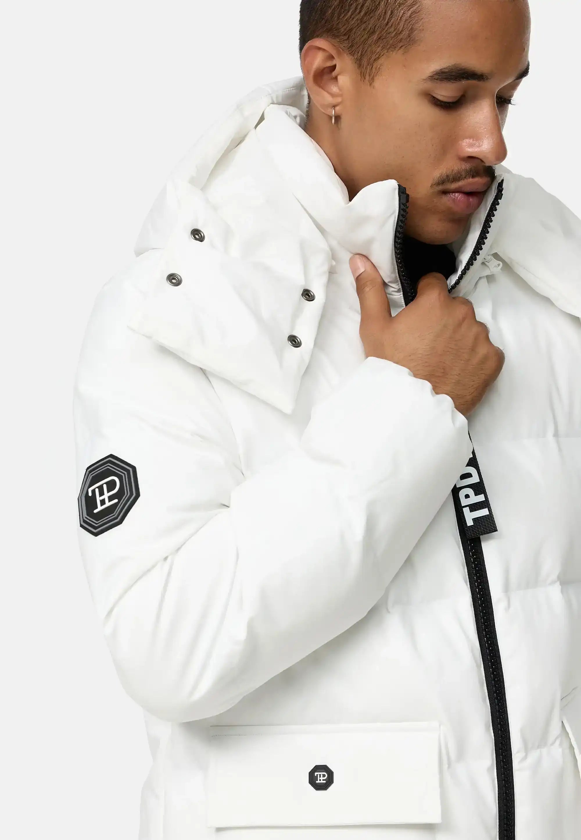 Eine Person in der Trueprodigy Franco Winterjacke mit abnehmbarer Kapuze, weiß mit schwarzen Reißverschlüssen und Logo-Patches an Ärmel und Tasche, steht vor hellem Hintergrund, blickt nach unten und hält den Kragen.