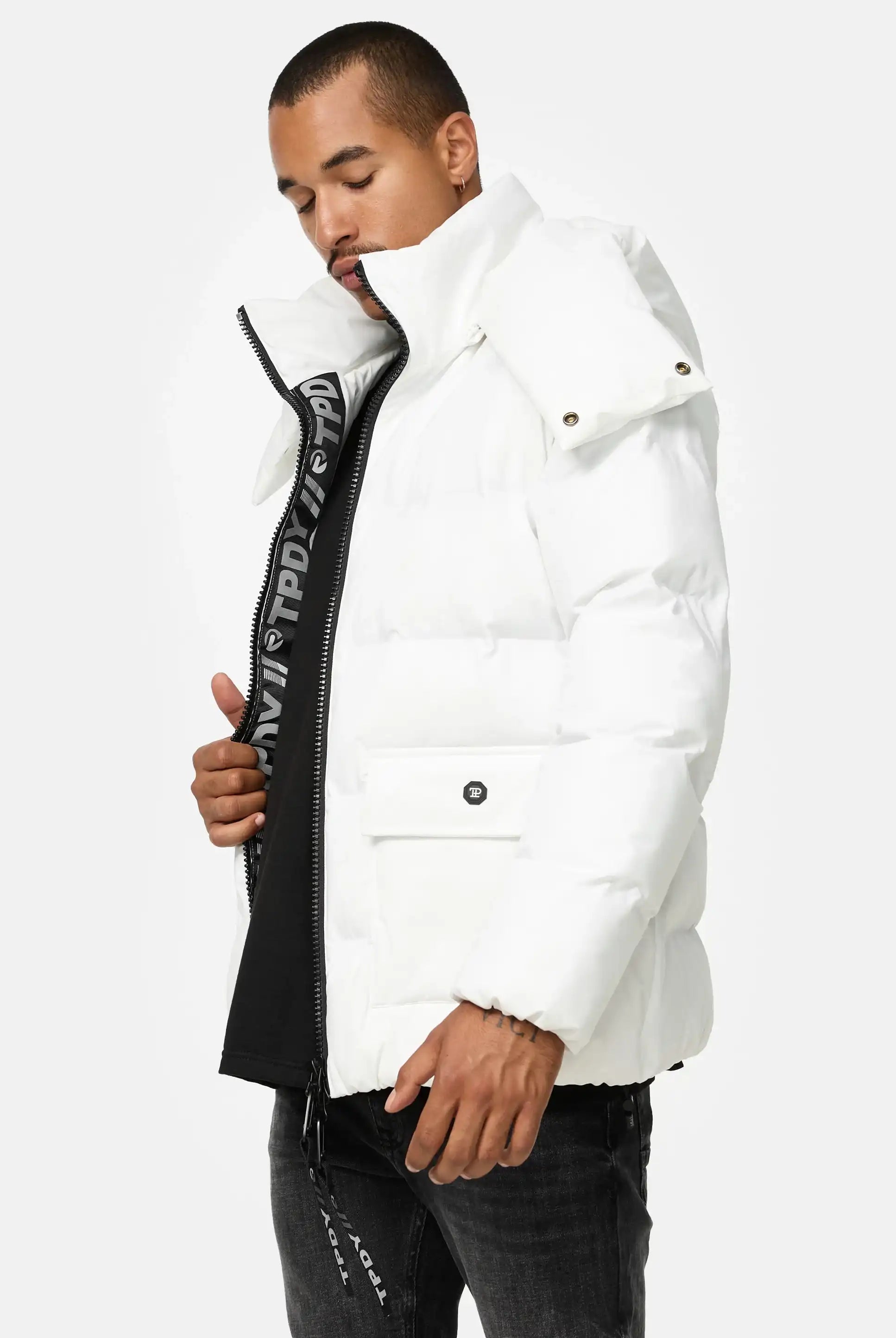 Ein Mann, der die Trueprodigy Franco Winterjacke mit abnehmbarer Kapuze, eine weiße Puffjacke mit hohem Kragen, trägt, steht vor einem schlichten Hintergrund. Er öffnet teilweise den Reißverschluss, so dass ein schwarzes Hemd darunter zum Vorschein kommt, und hat die Augen geschlossen.