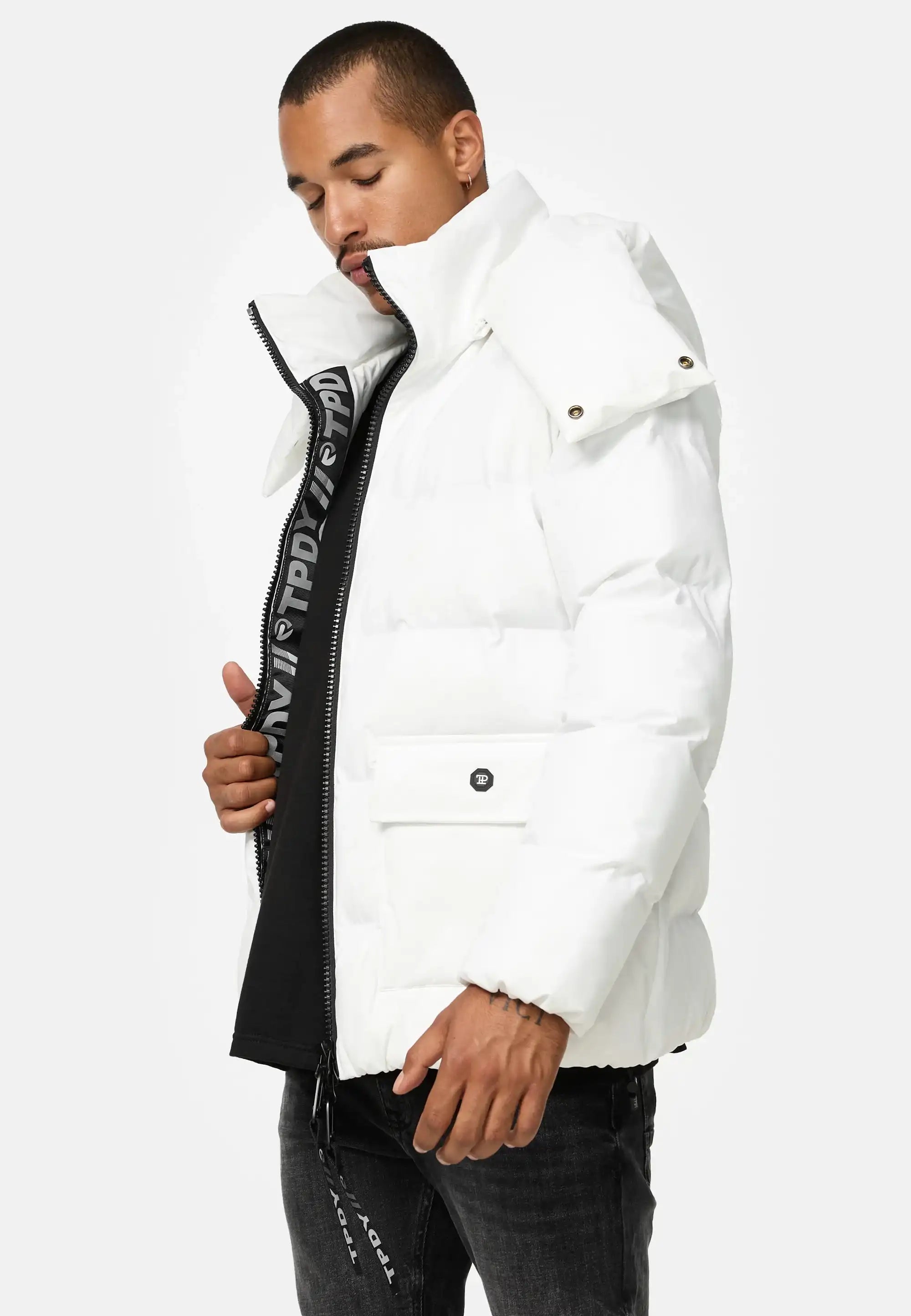 Ein Mann, der die Trueprodigy Franco Winterjacke mit abnehmbarer Kapuze, eine weiße Puffjacke mit hohem Kragen, trägt, steht vor einem schlichten Hintergrund. Er öffnet teilweise den Reißverschluss, so dass ein schwarzes Hemd darunter zum Vorschein kommt, und hat die Augen geschlossen.