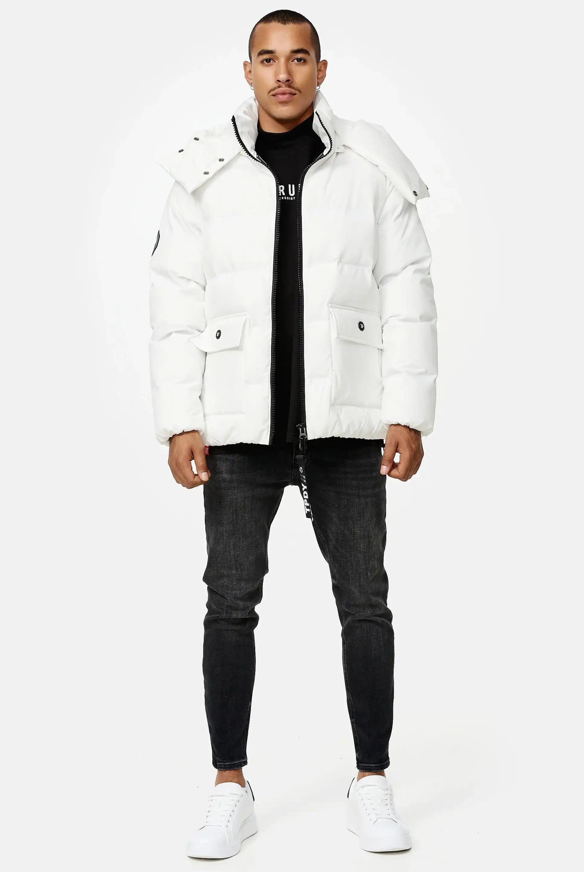 Ein Mann sieht nach vorne in einer Trueprodigy Franco Winterjacke mit abnehmbarer Kapuze, schwarzem Hemd, dunkler Skinny Jeans und weißen Turnschuhen vor einem schlichten hellen Hintergrund.