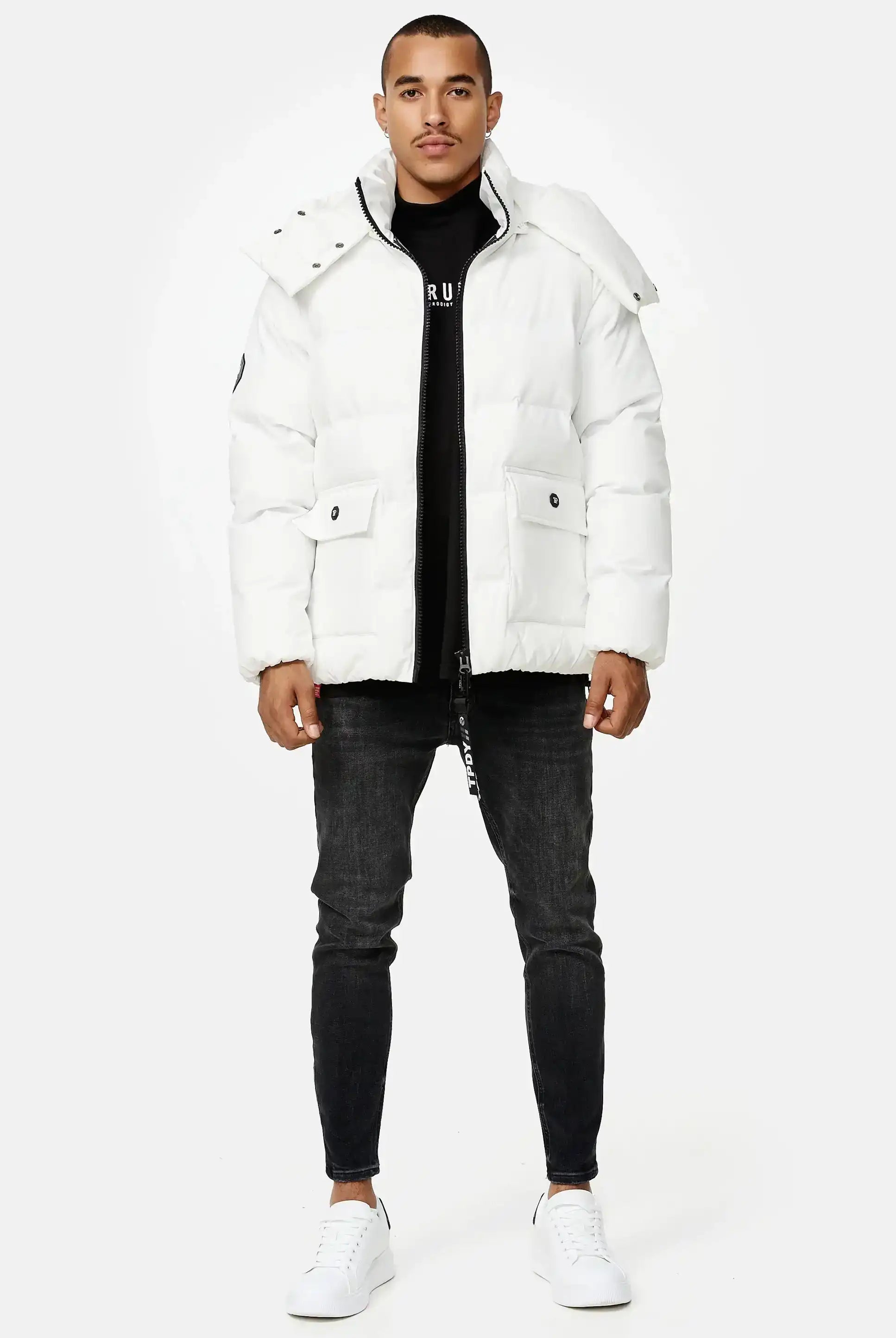 Ein Mann sieht nach vorne in einer Trueprodigy Franco Winterjacke mit abnehmbarer Kapuze, schwarzem Hemd, dunkler Skinny Jeans und weißen Turnschuhen vor einem schlichten hellen Hintergrund.
