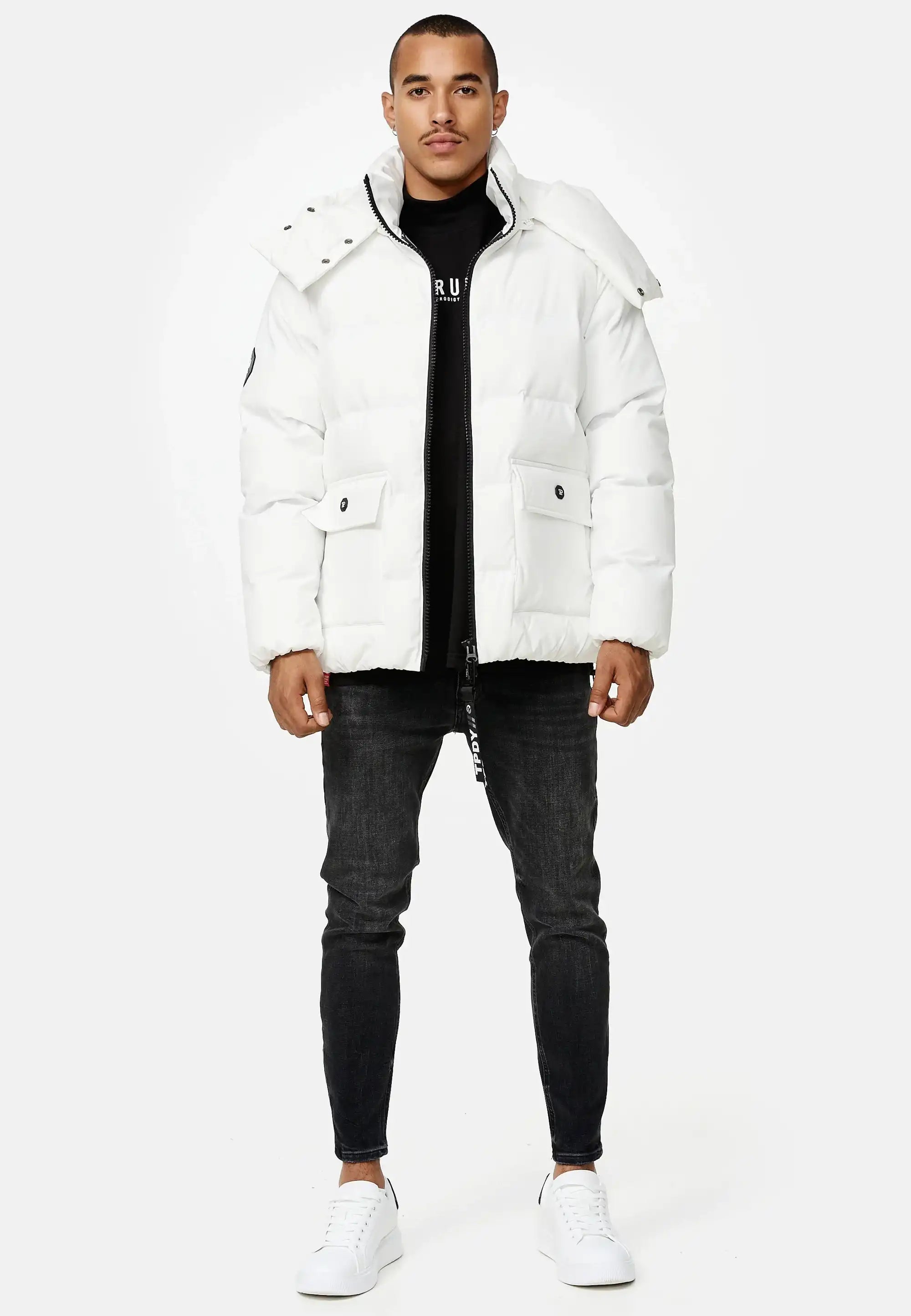 Ein Mann sieht nach vorne in einer Trueprodigy Franco Winterjacke mit abnehmbarer Kapuze, schwarzem Hemd, dunkler Skinny Jeans und weißen Turnschuhen vor einem schlichten hellen Hintergrund.