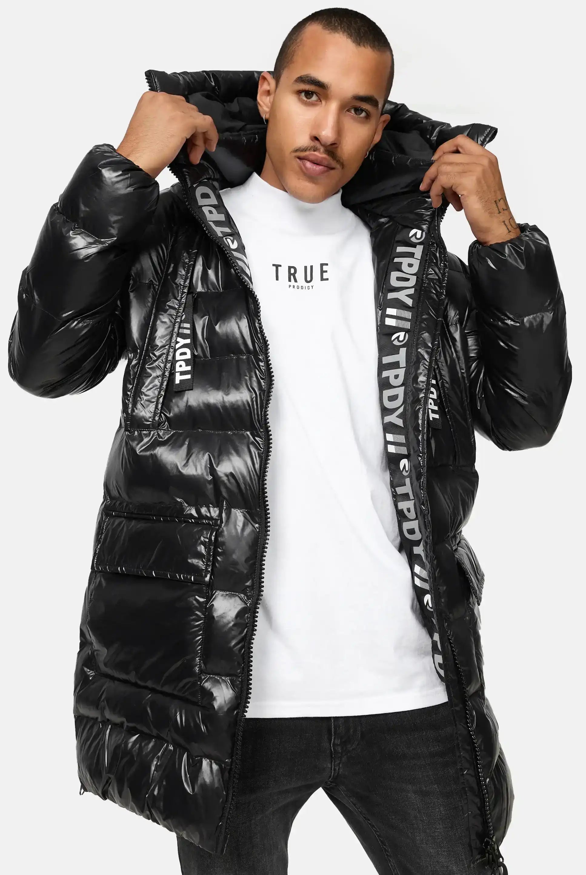 Ein Mann trägt die David Glänzende Steppjacke mit Kapuze von trueprodigy über einem weißen TRUE-T-Shirt und einer schwarzen Jeans und steht mit den Händen an den Seiten der Kapuze.