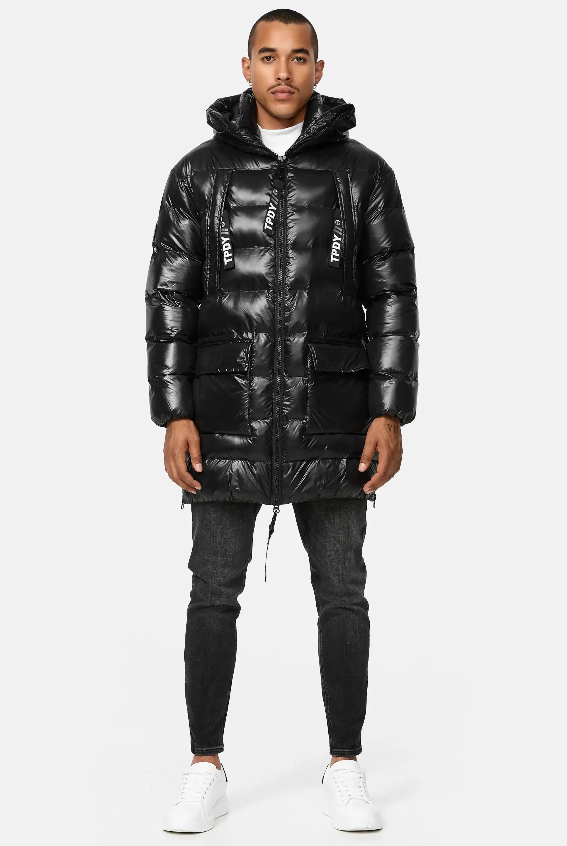 Ein Mann steht mit dem Gesicht nach vorne und trägt die trueprodigy David Glänzende Steppjacke mit Kapuze mit großen Vordertaschen und Text auf den Kordelzügen, gepaart mit dunklen Jeans und weißen Turnschuhen vor einem schlichten hellen Hintergrund.