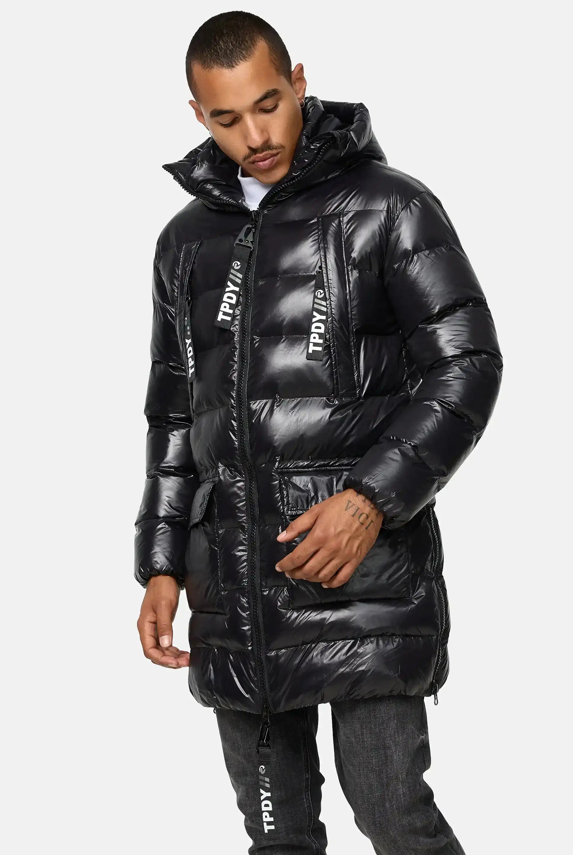 Eine Person, die die trueprodigy David Glänzende Steppjacke mit Kapuze mit schwarzem Finish und großen Vordertaschen trägt, gepaart mit einer dunklen Jeans, steht vor einem schlichten hellgrauen Hintergrund.