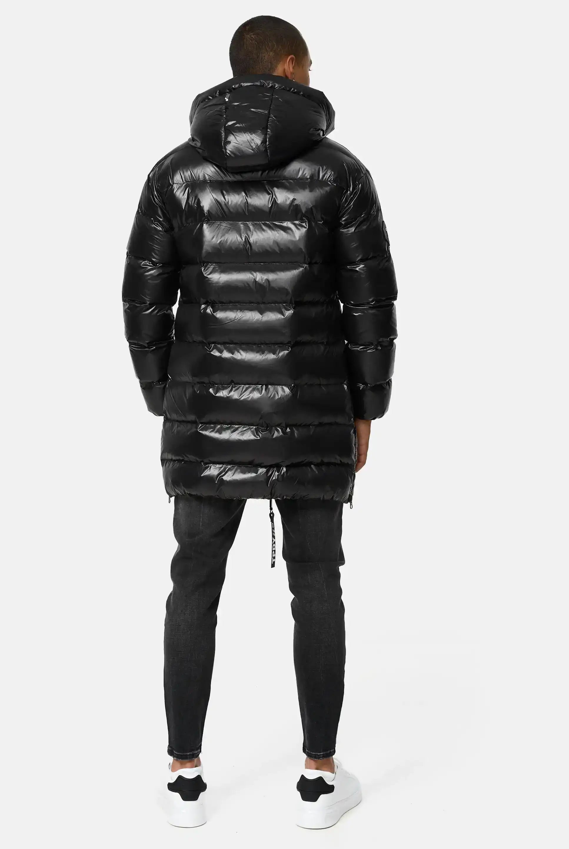 Eine Person steht mit dem Gesicht zur Wand und trägt die trueprodigy David Glänzende Steppjacke mit Kapuze, dunkle Skinny Jeans und weiße Turnschuhe. Der Hintergrund ist einfarbig und hell.