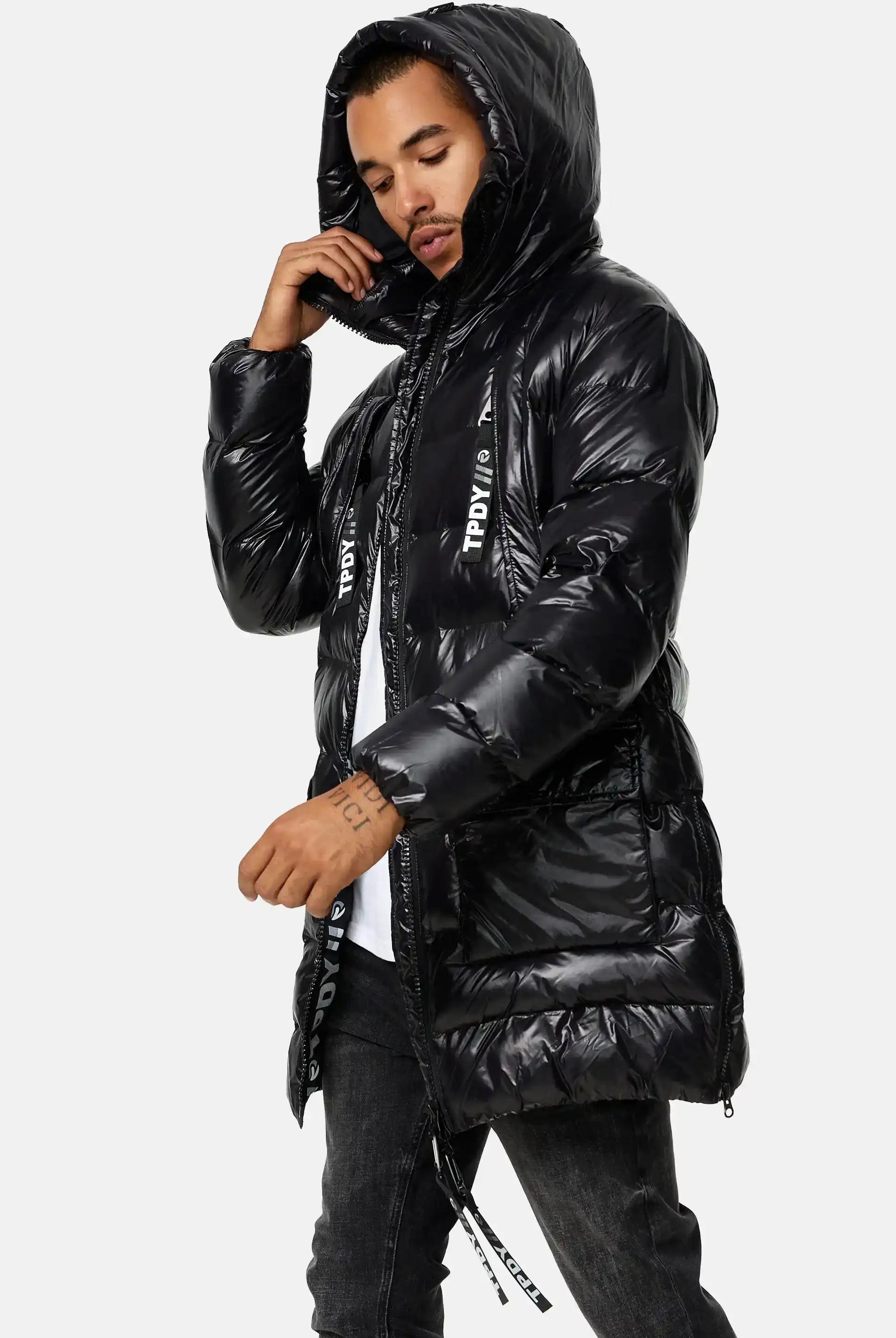 Eine Person in einer Trueprodigy David Glänzende Steppjacke mit Kapuze mit Reißverschlüssen und Kordelzügen vorne, gepaart mit einer dunklen Jeans, steht vor einem schlichten hellen Hintergrund.