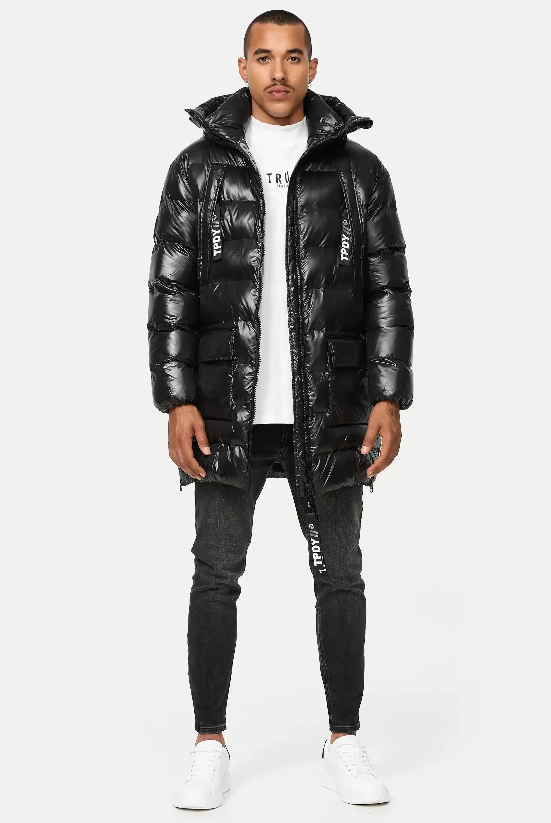 Ein Mann blickt nach vorne und trägt die Trueprodigy David Glänzende Steppjacke mit Kapuze über einem weißen T-Shirt, schwarze Jeans und weiße Turnschuhe. Der Hintergrund ist einfarbig weiß.