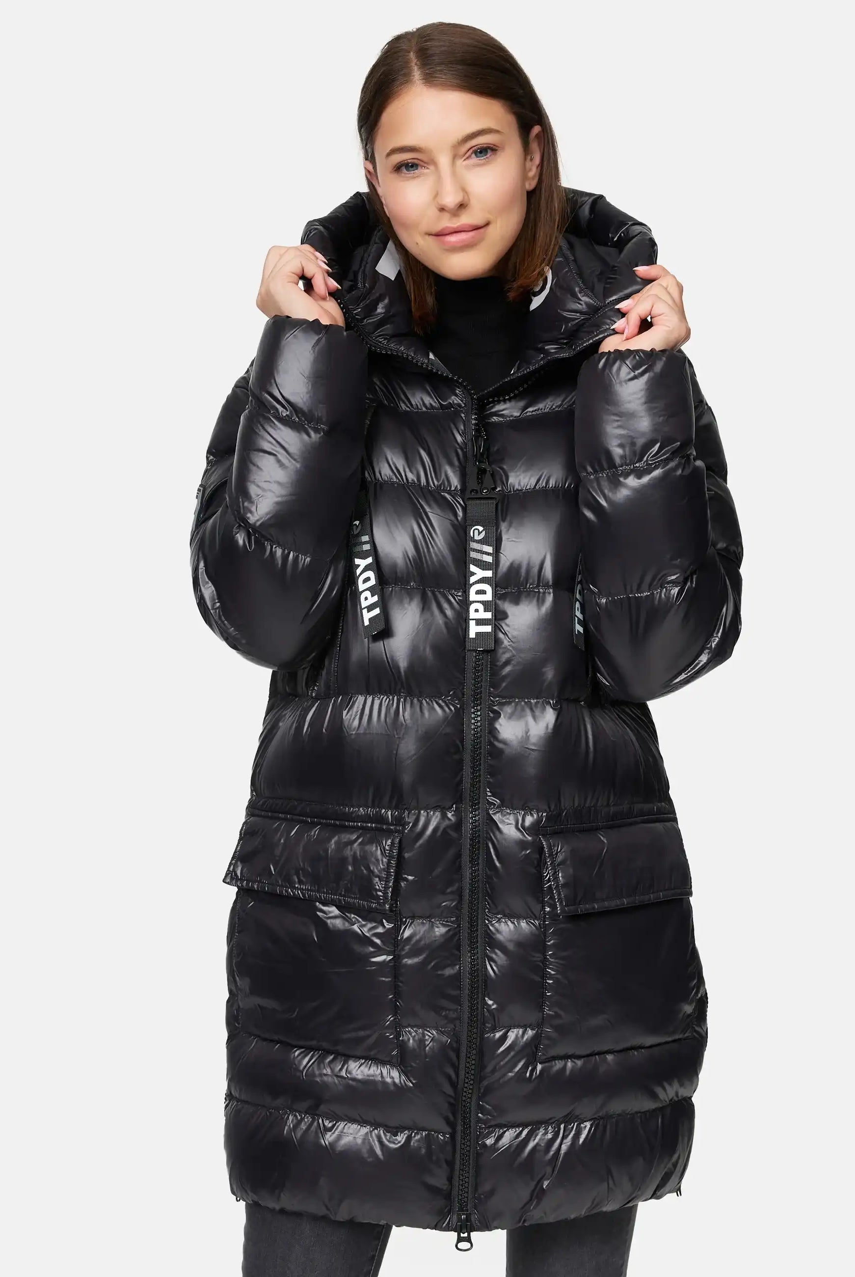 Eine Frau, die die Trueprodigy David F Glänzende Steppjacke mit Kapuze trägt, steht mit dem Gesicht nach vorne, hält die Kapuze mit beiden Händen und lächelt sanft.