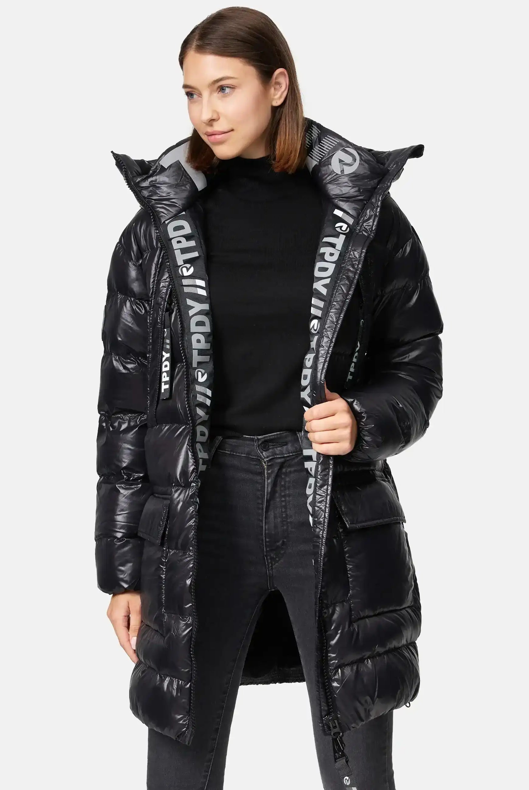 Eine Frau trägt die trueprodigy David F Glänzende Steppjacke mit Kapuze mit hohem Kragen über einem schwarzen Rollkragen und dunklen Jeans. Das Innenfutter der Jacke ist mit weißer Schrift versehen, während sie leicht zur Seite gewandt steht.