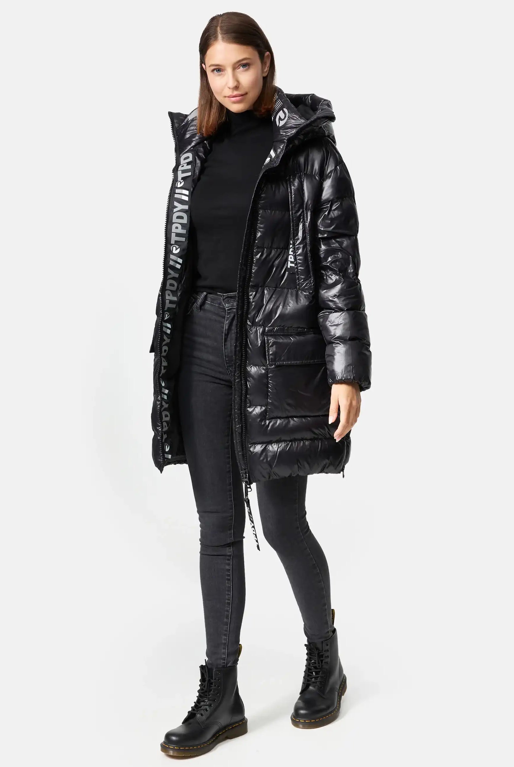 Eine Frau blickt nach vorne und trägt die trueprodigy David F Glänzende Steppjacke mit Kapuze mit großen Taschen auf der Vorderseite und Text auf der Innenkapuze. Dazu trägt sie einen schwarzen Rollkragenpullover, graue Skinny Jeans und schwarze Schnürstiefel.