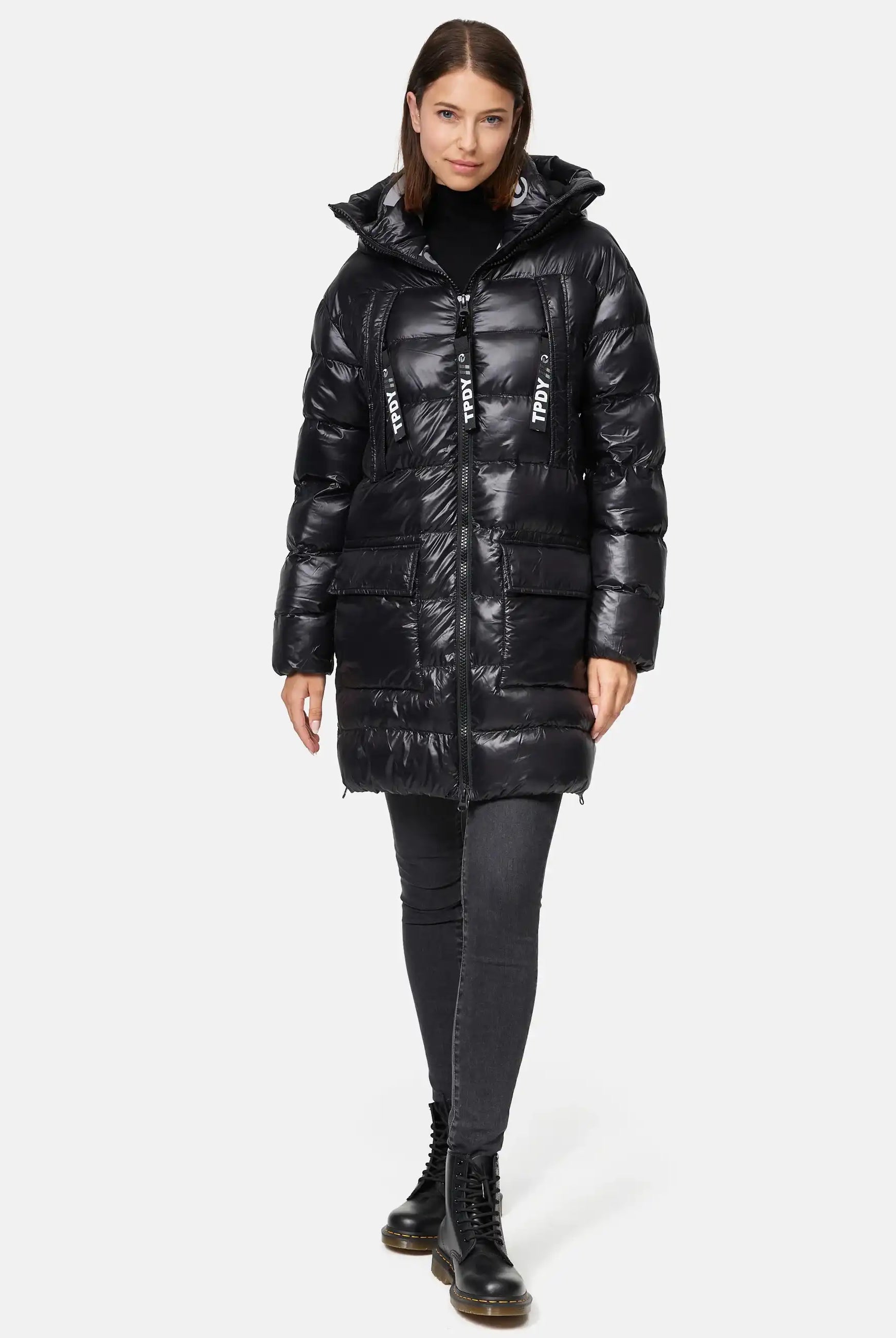 Eine Frau blickt nach vorne in der trueprodigy David F Glänzende Steppjacke mit Kapuze, einem glänzenden schwarzen Puff mit Kapuze, großen Reißverschlüssen mit weißem Text, gepaart mit schwarzen Jeans und Schnürstiefeln. Ihr Haar ist glatt und schulterlang.