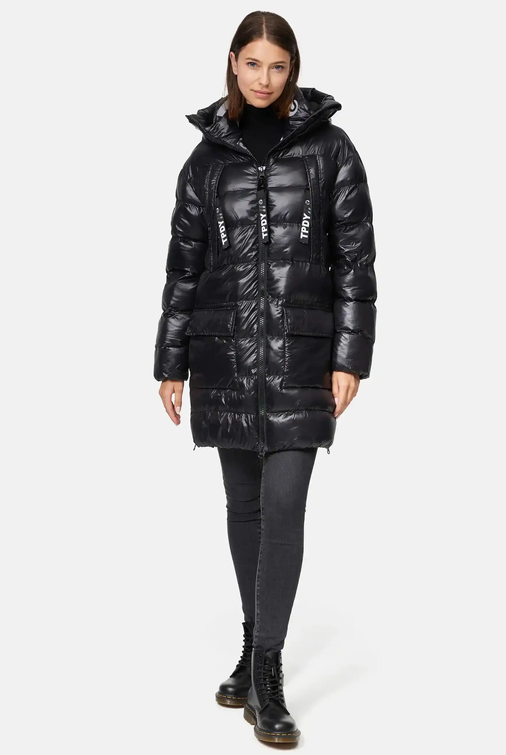 Eine Frau blickt nach vorne in der trueprodigy David F Glänzende Steppjacke mit Kapuze, einem glänzenden schwarzen Puff mit Kapuze, großen Reißverschlüssen mit weißem Text, gepaart mit schwarzen Jeans und Schnürstiefeln. Ihr Haar ist glatt und schulterlang.