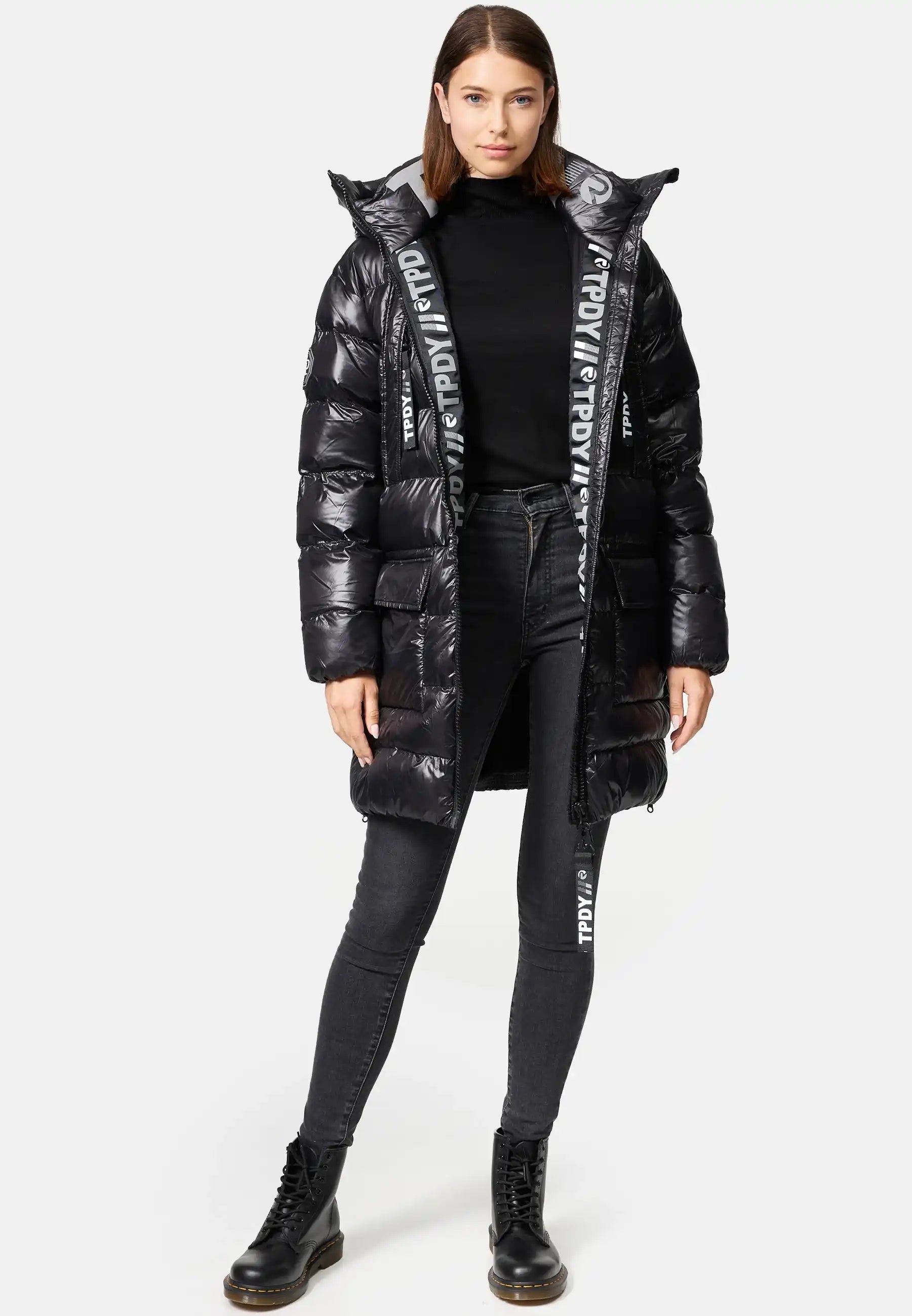 Eine Frau blickt nach vorne und trägt die trueprodigy David F Glänzende Steppjacke mit Kapuze mit bedrucktem Innenfutter, gepaart mit einem schwarzen Rollkragenpullover, schwarzen Skinny Jeans und schwarzen Schnürstiefeln vor einem schlichten hellen Hintergrund.