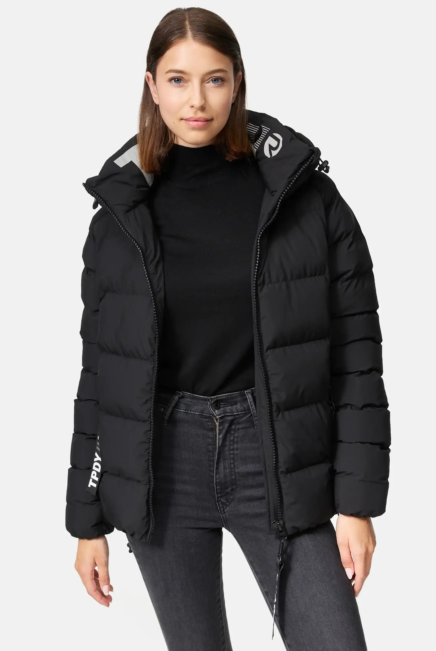 Eine Frau trägt die trueprodigy Murphy F Matte Steppjacke mit Kapuze in Schwarz, offen über einem schwarzen Rollkragenpullover und grauen Jeans. Die wasserabweisende Jacke mit horizontalem Steppmuster hat einen schlichten weißen Hintergrund.