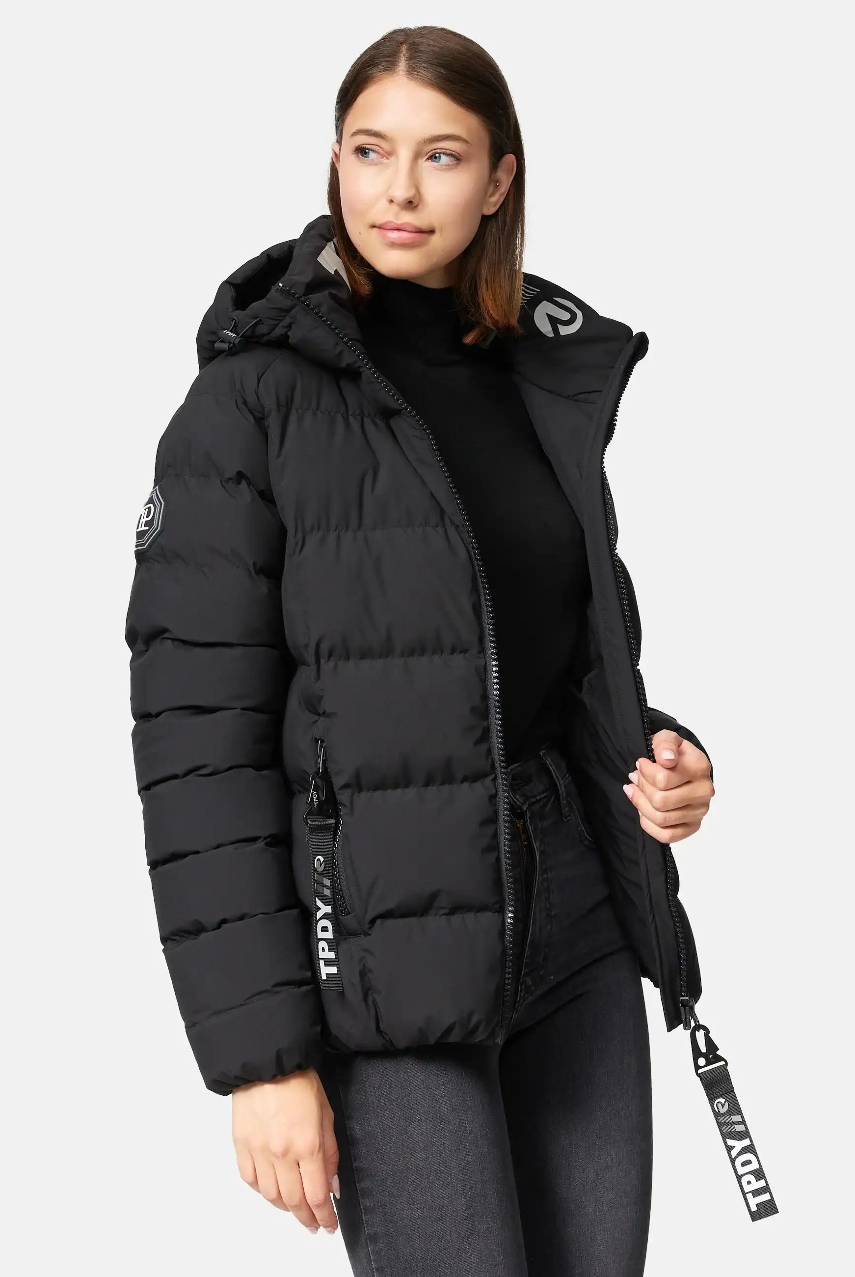 Eine Frau trägt die schwarze Murphy F Matte Steppjacke mit Kapuze von trueprodigy, wasserabweisend mit sichtbaren TPDY-Zippern und rundem Ärmel-Aufnäher, dazu einen schwarzen Rollkragen und dunkle Jeans vor hellem Hintergrund.